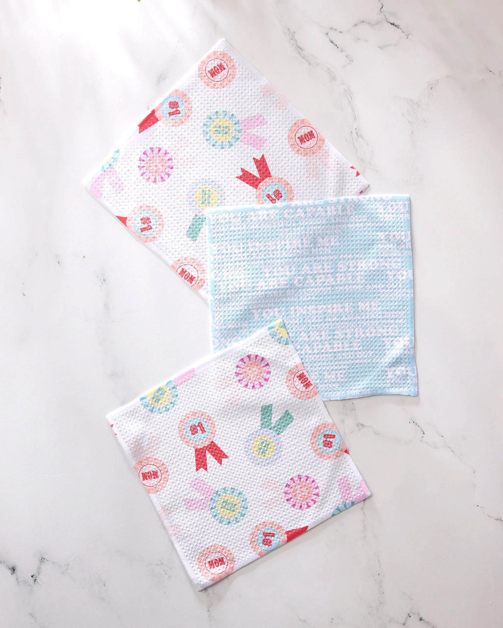 Three GEOMETRY Mom Mantra Swedish dishcloths, made from recycled materials, rest on marble; two showcase ribbon and medal patterns, while one is light blue with white text—an eco-friendly, super-absorbent kitchen set.
