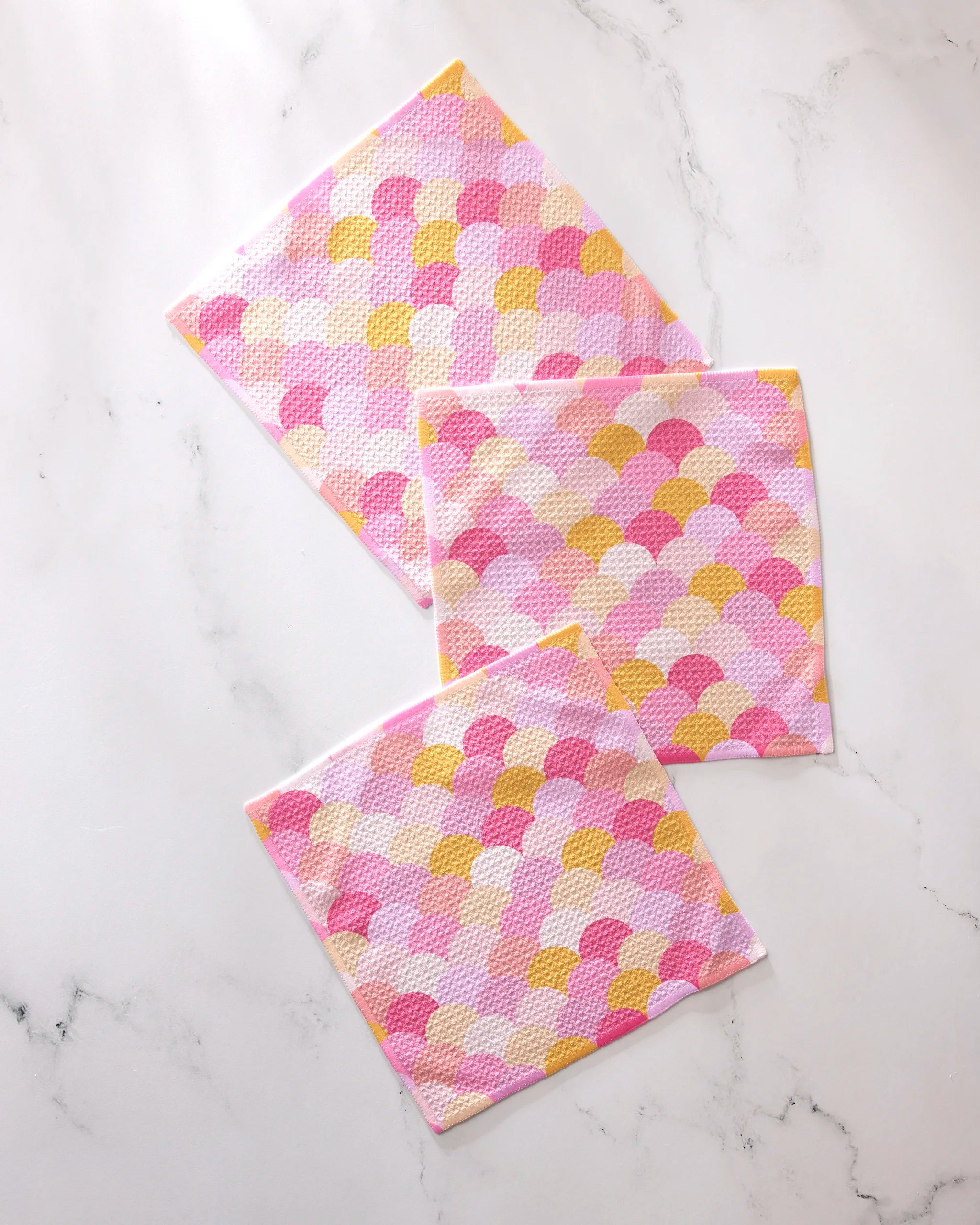The GEOMETRY Inherit dishcloth set includes three super-absorbent towels in pink, yellow, and white scallop patterns, artfully displayed on a white marble surface.