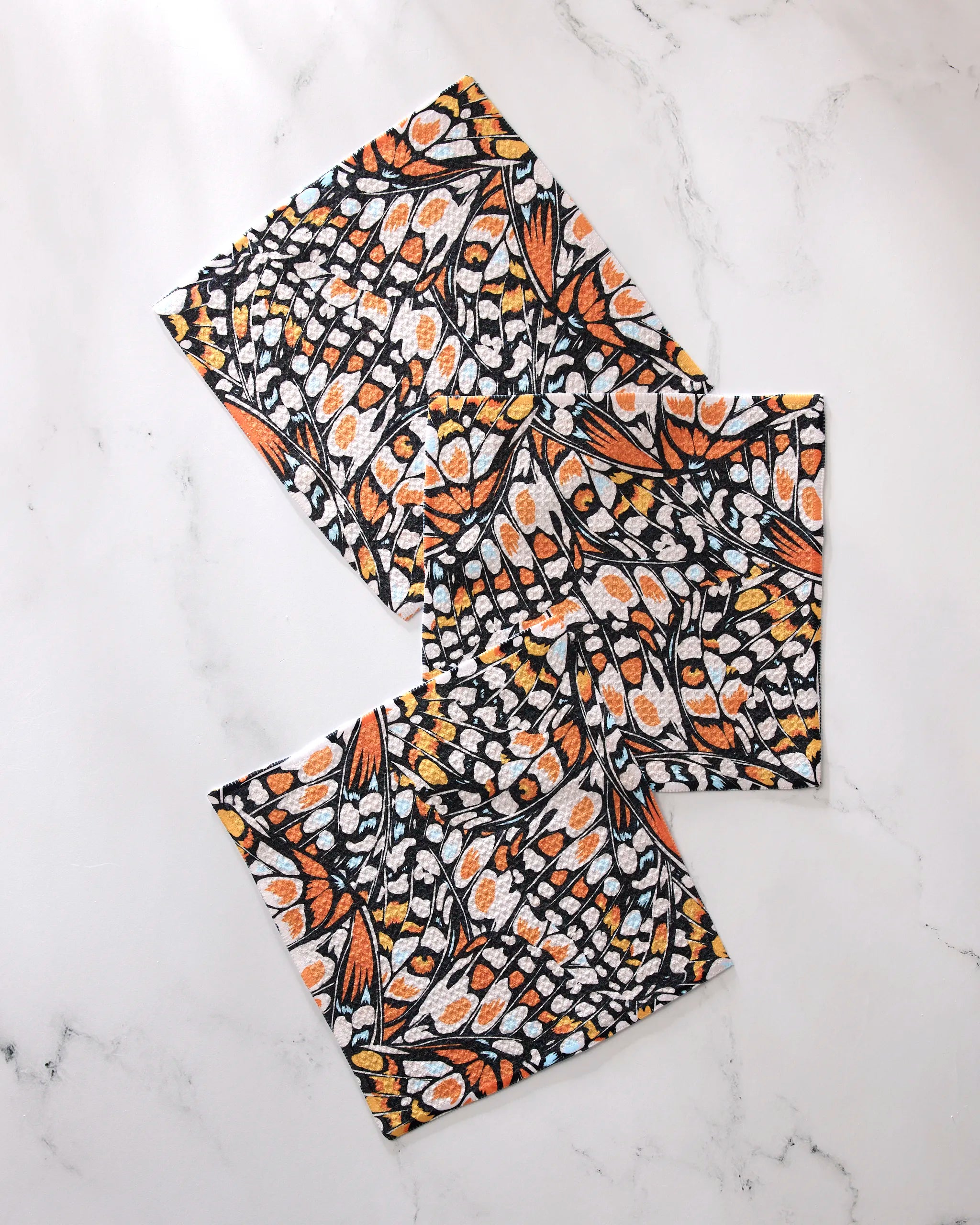 Three "Wings in the Wind" fabric napkins by GEOMETRY, featuring a vibrant monarch butterfly wing pattern in orange, black, yellow, and white recycled material, are arranged in a scattered layout on a white marble surface.