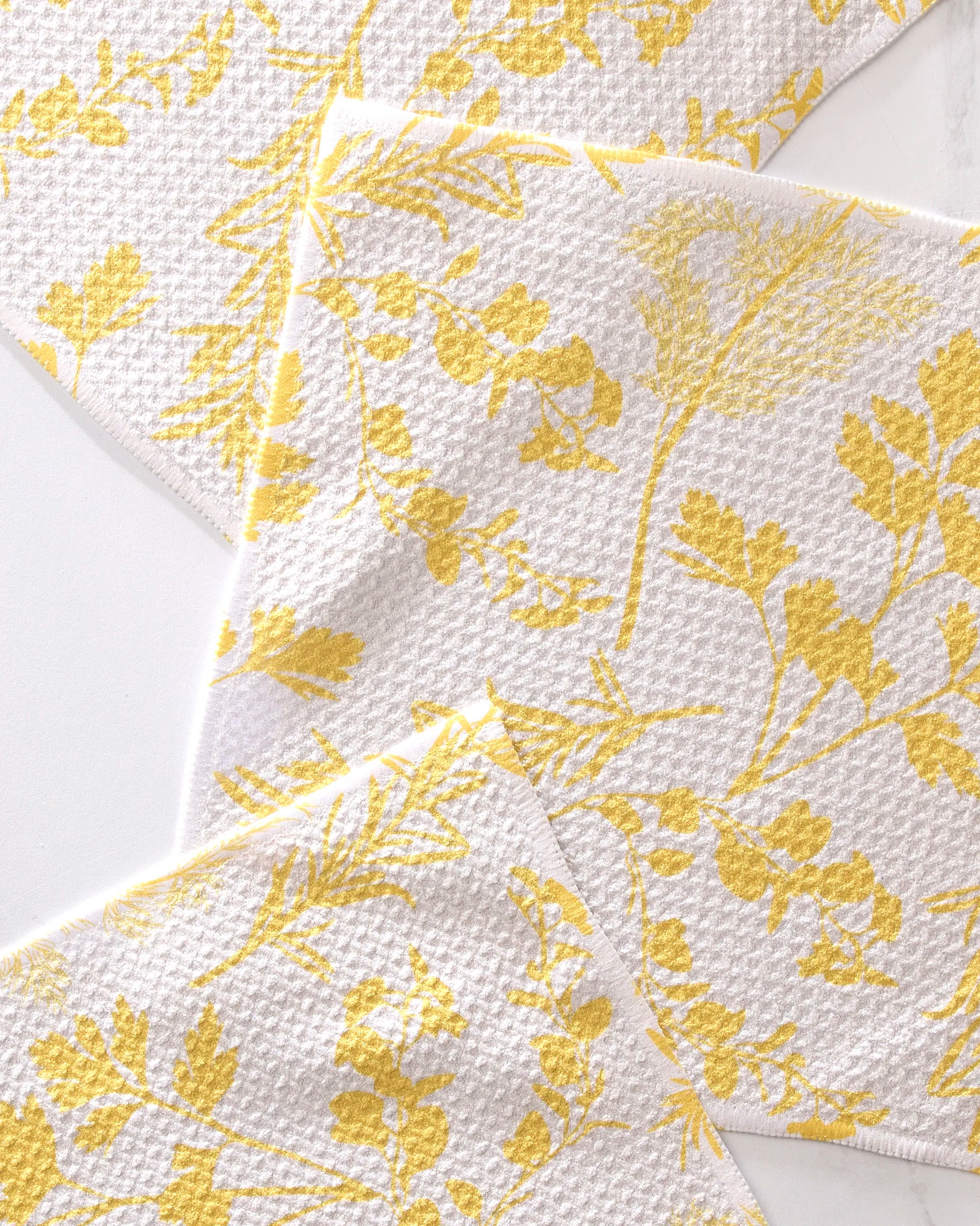 Three "Garden of Gold" napkins by GEOMETRY, made from recycled materials with textured surfaces and yellow botanical prints, overlap on a white background, creating a bright, cheerful display.