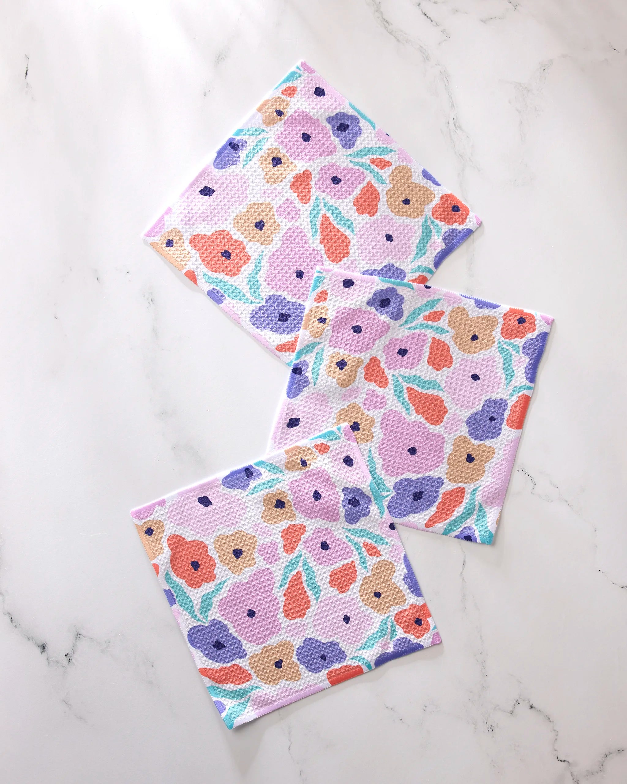 The GEOMETRY Blossoming Love set features three square dishcloths with colorful orange, purple, pink, and beige floral patterns with green leaves, arranged on a marble-like surface. Made from recycled materials for vibrant style.