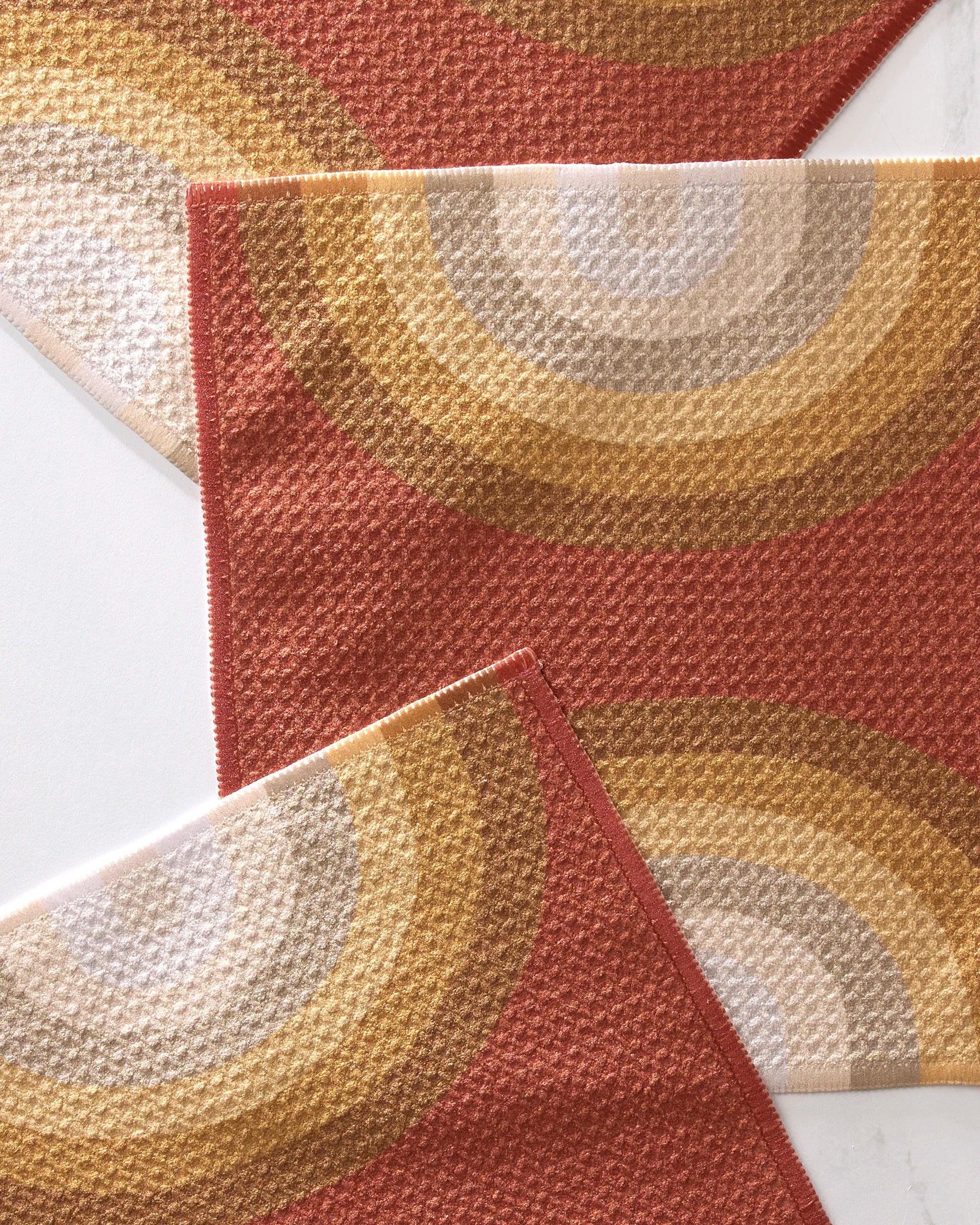 Three "Pumpkin Spice" placemats by GEOMETRY, made from recycled materials, feature a retro semicircle pattern in gold, beige, and white on a rust background. Their textured fabric and overlapping edges are displayed on a light surface.