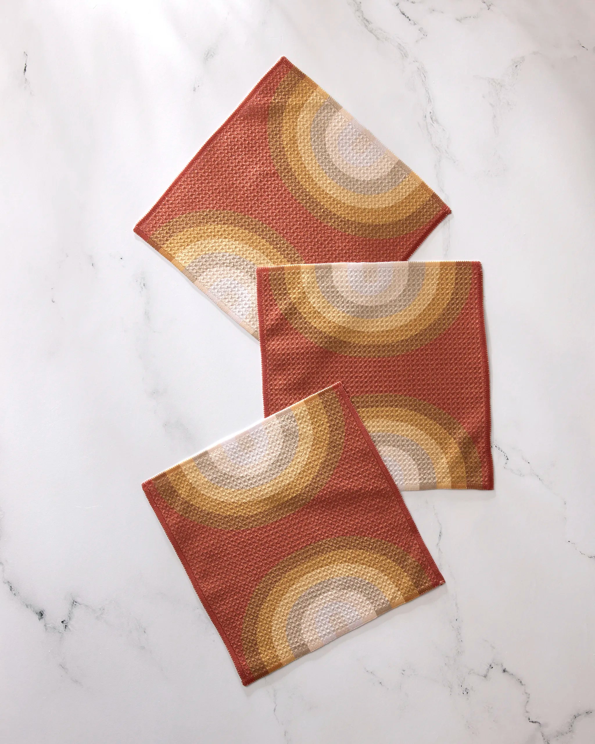 Three "Pumpkin Spice" square cloth napkins by GEOMETRY, made from recycled materials with a retro orange, yellow, and beige circular pattern, are displayed on a white marble surface.