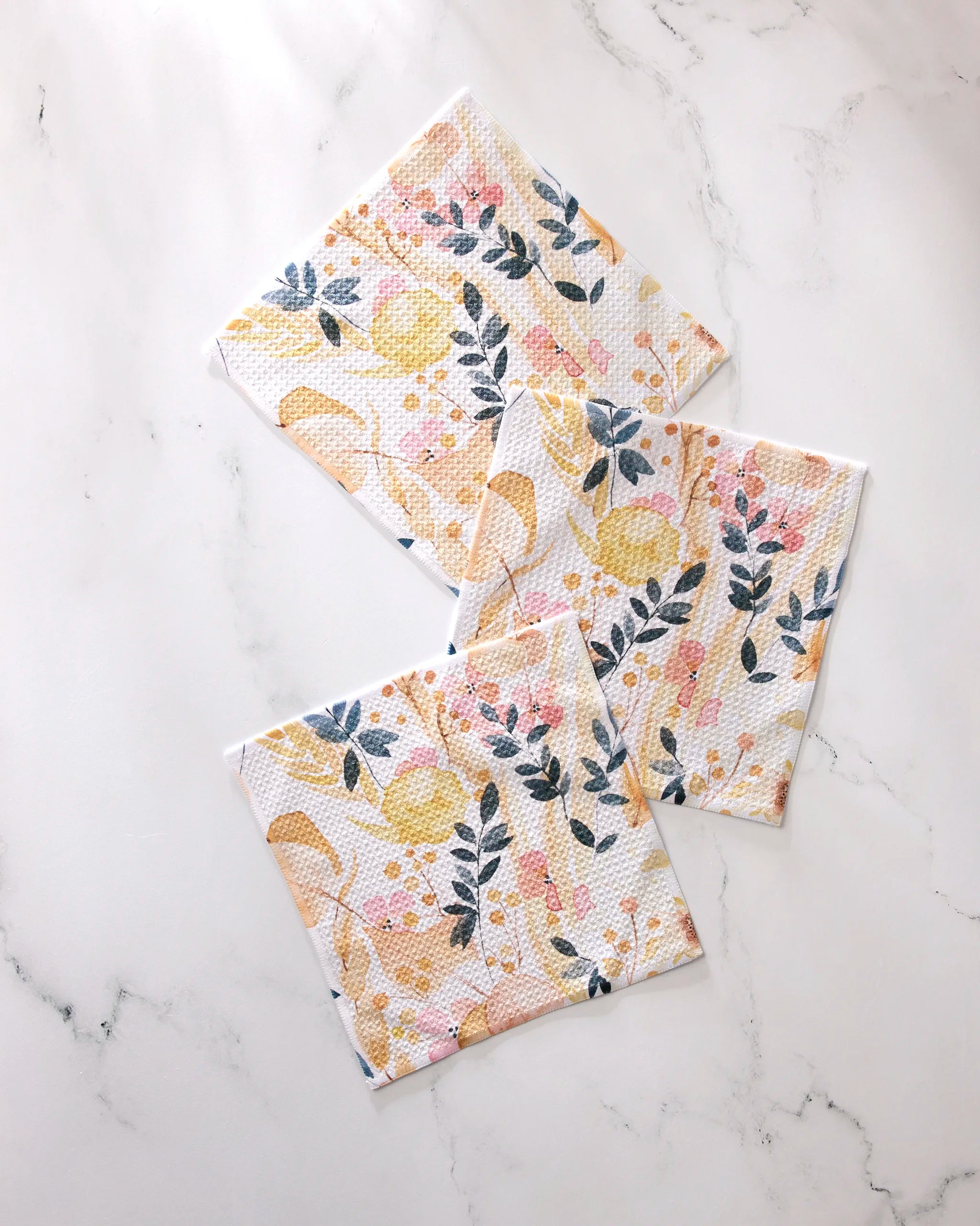 Three "Sleepy Fields" paper napkins by GEOMETRY, featuring yellow, pink, and blue floral patterns, are displayed on a white marble surface—an elegant addition to any dishcloth set.