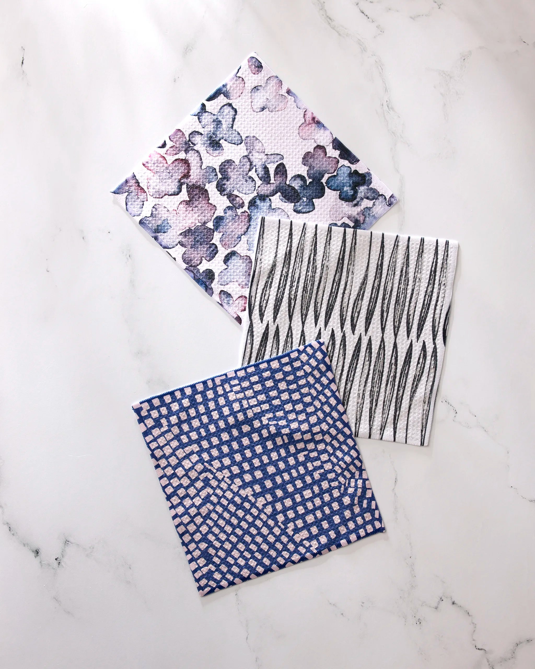 Three GEOMETRY Tiny Petals dishcloths—featuring purple and blue florals, black wavy lines on white, and a blue-white grid—are made from super-absorbent recycled materials and displayed on a white marble surface.