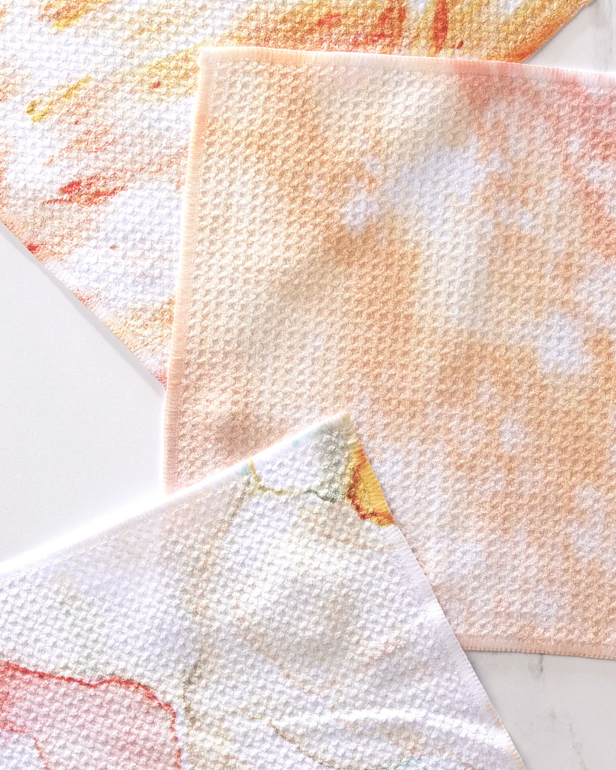 The GEOMETRY Nice Water Dishcloth Set includes three super-absorbent cloths with abstract pastel designs in orange, peach, yellow, and white. Made from recycled materials, each features a subtle waffle texture.