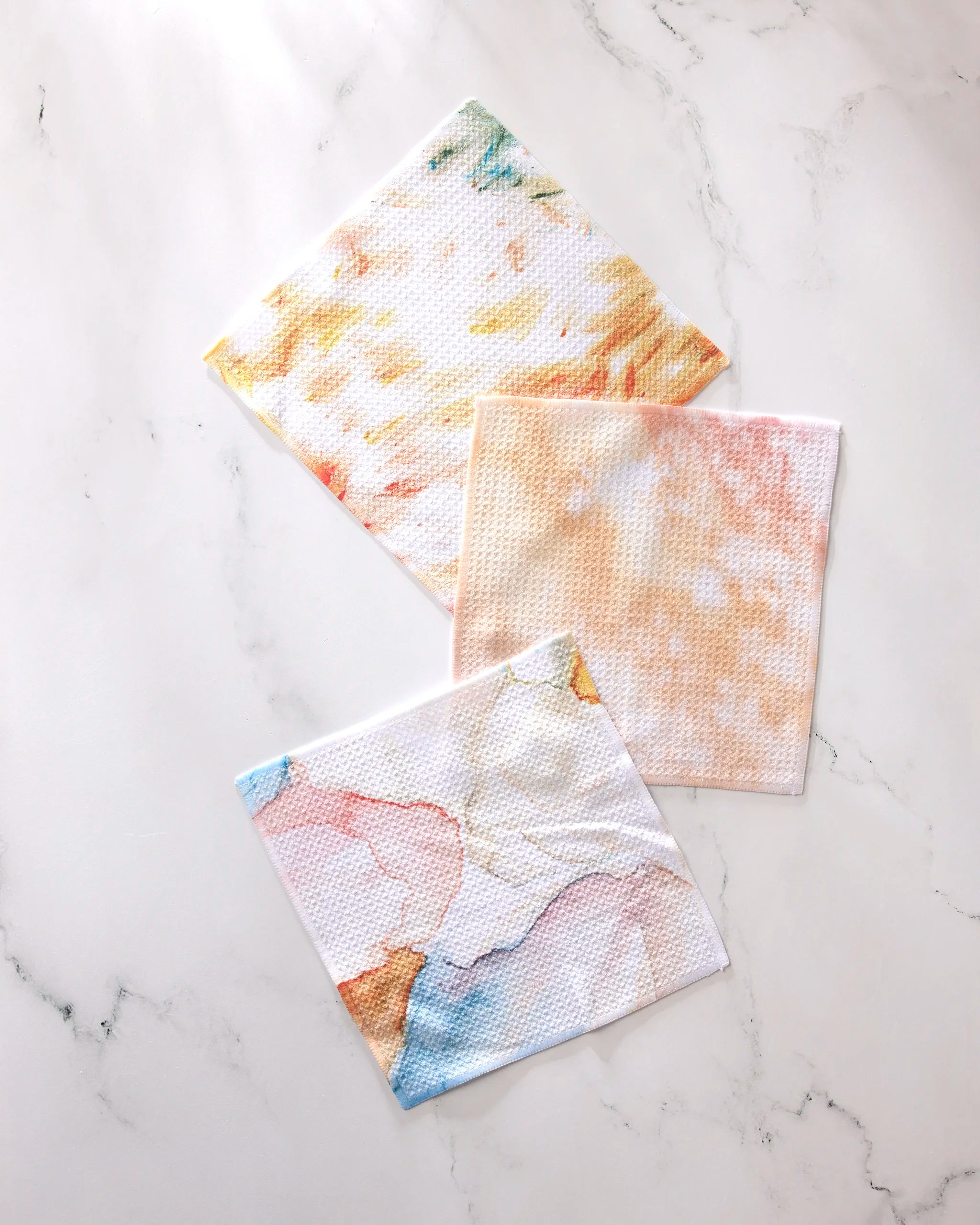 Three square cloths from GEOMETRY's Nice Water collection—featuring pastel marble patterns in yellow, orange, and multicolor—are displayed on white marble. Crafted from recycled materials, these super-absorbent dishtowels are both stylish and eco-friendly.