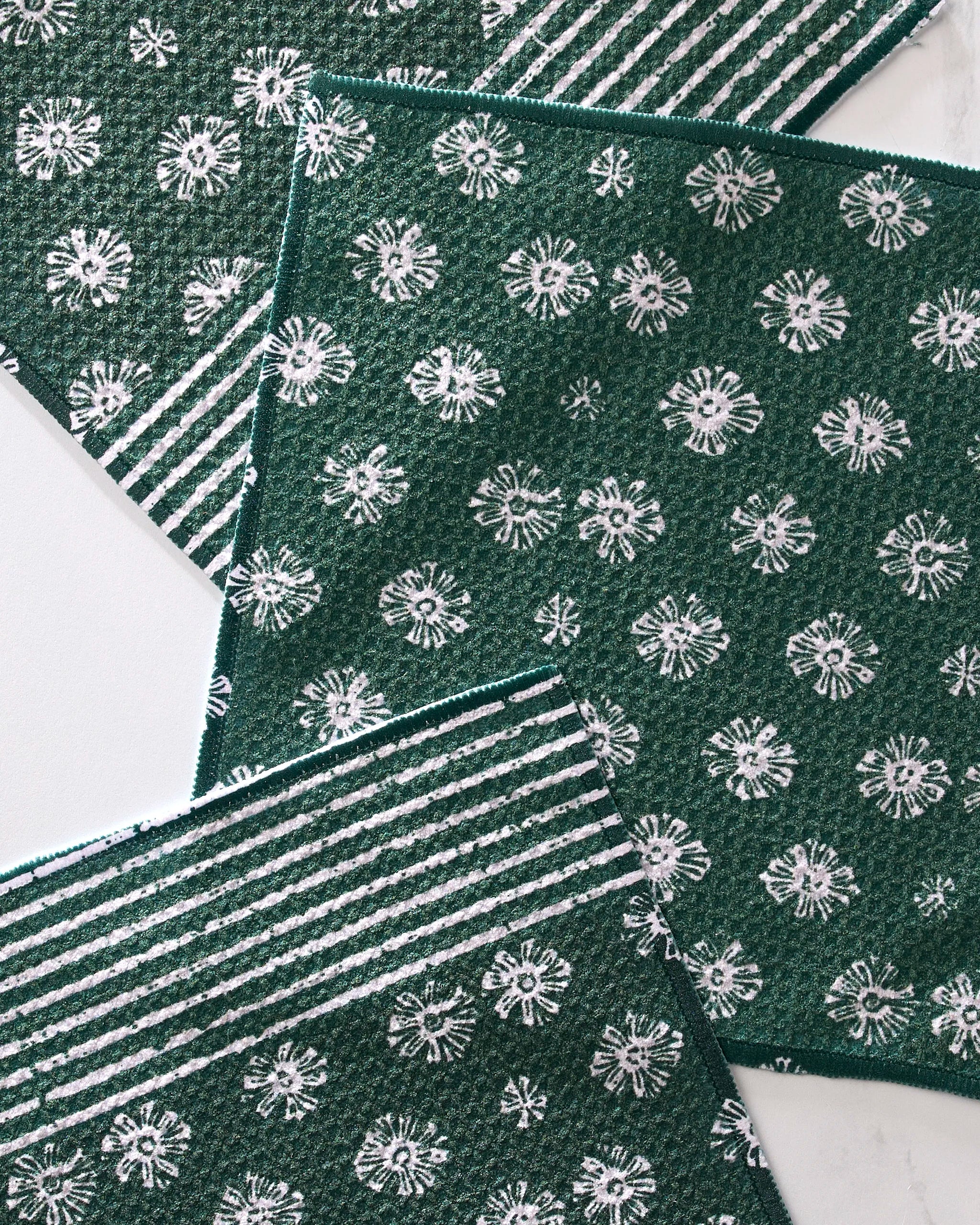 Three green Arbusto kitchen towels by Geometry are arranged overlapping on a white surface; two have white floral patterns and one displays white stripes.