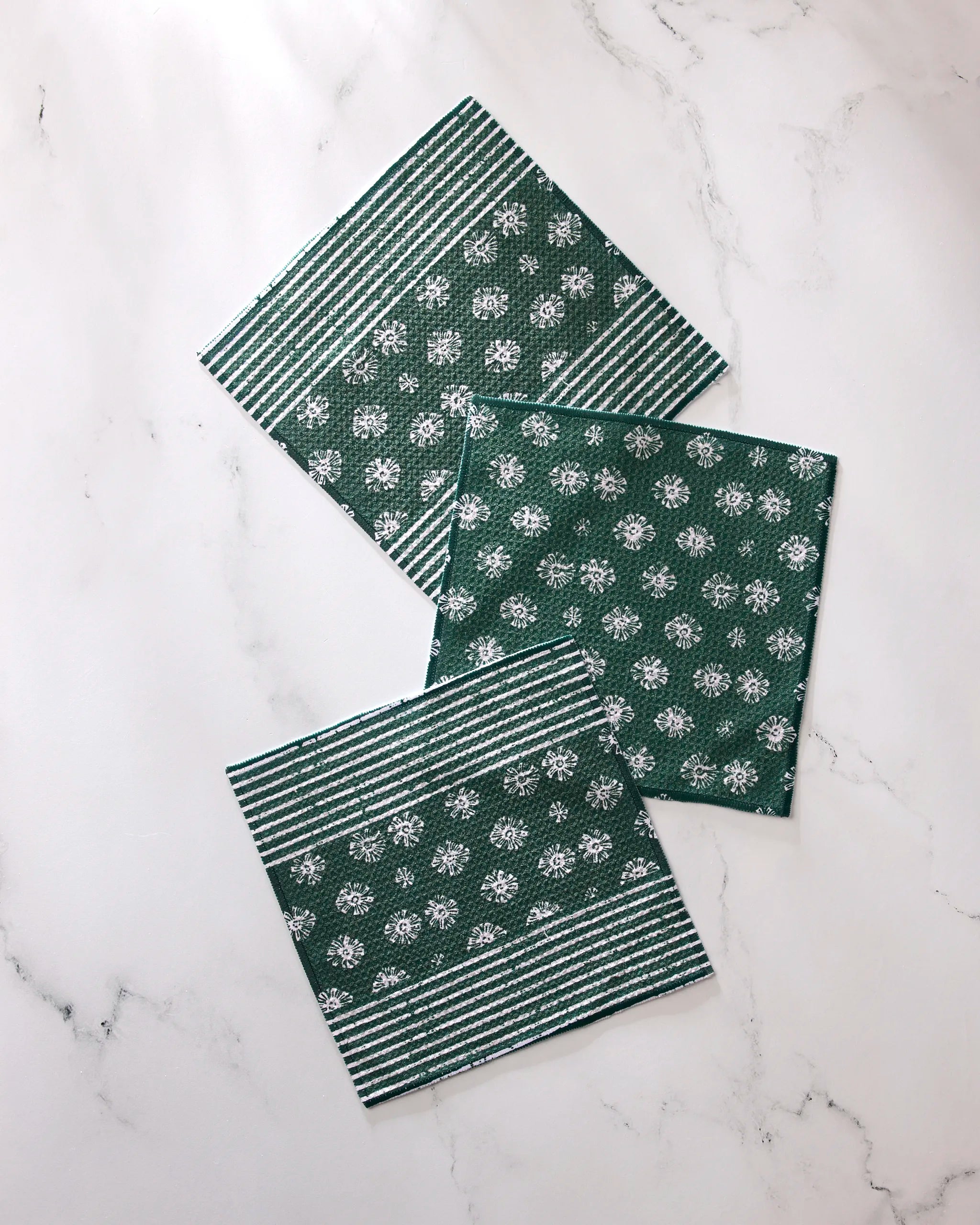Three green Geometry Arbusto absorbent kitchen towels with white floral and stripe patterns are arranged on a white marble surface—two feature both stripes and flowers, while one displays only the floral design.
