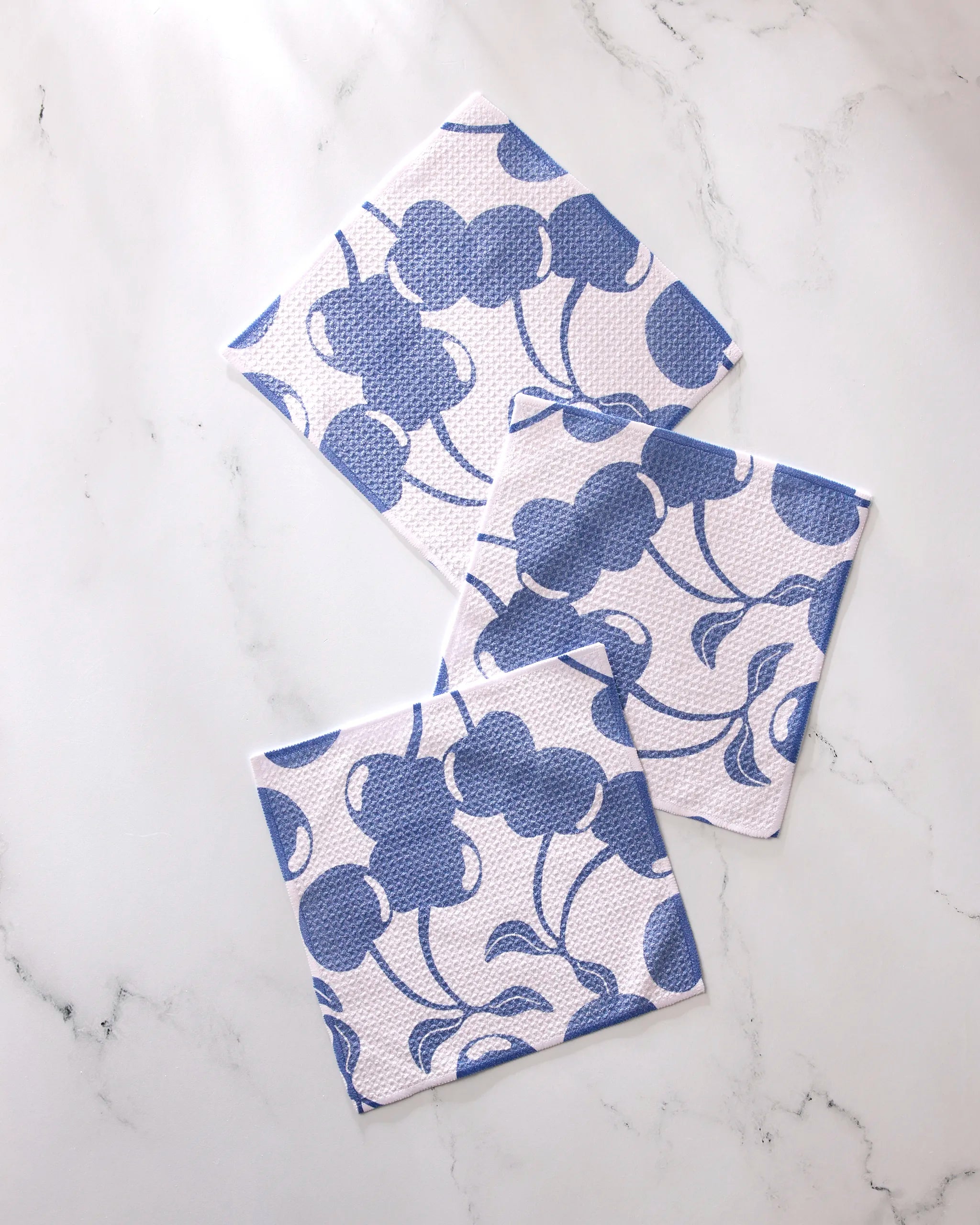 Three Geometry "Blue Cherry Dreams" napkins with a blue floral pattern are arranged on a white marble surface, slightly overlapping at different angles.