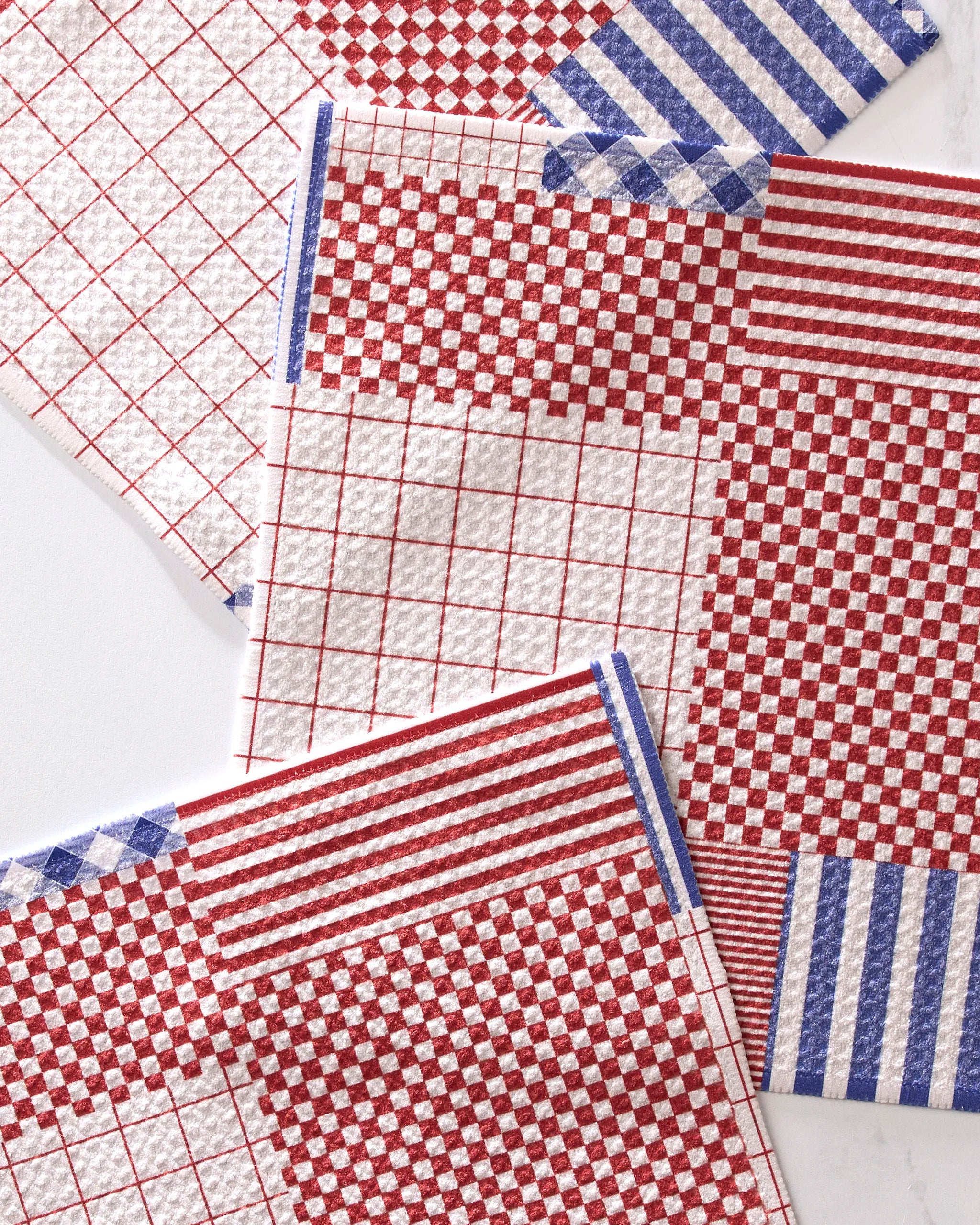 The Patched Up Parade dishcloth set by Geometry includes four super-absorbent dishtowels with red and blue geometric stripes, checks, and grids on a white background.