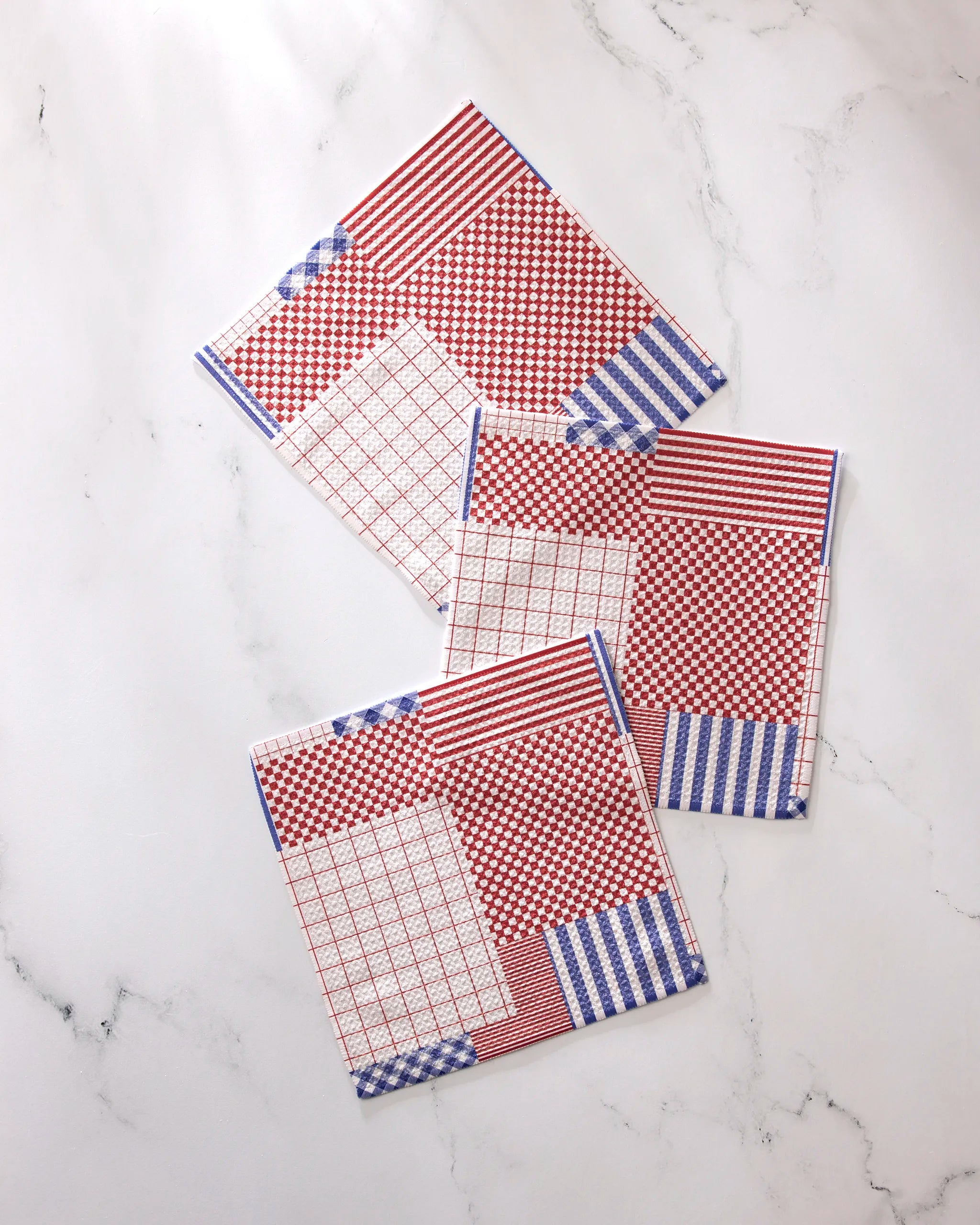 Three square napkins from Geometry’s Patched Up Parade set, featuring red, white, and blue geometric patterns—stripes, checks, and grids—are displayed on white marble. Made with recycled materials for an eco-friendly touch.