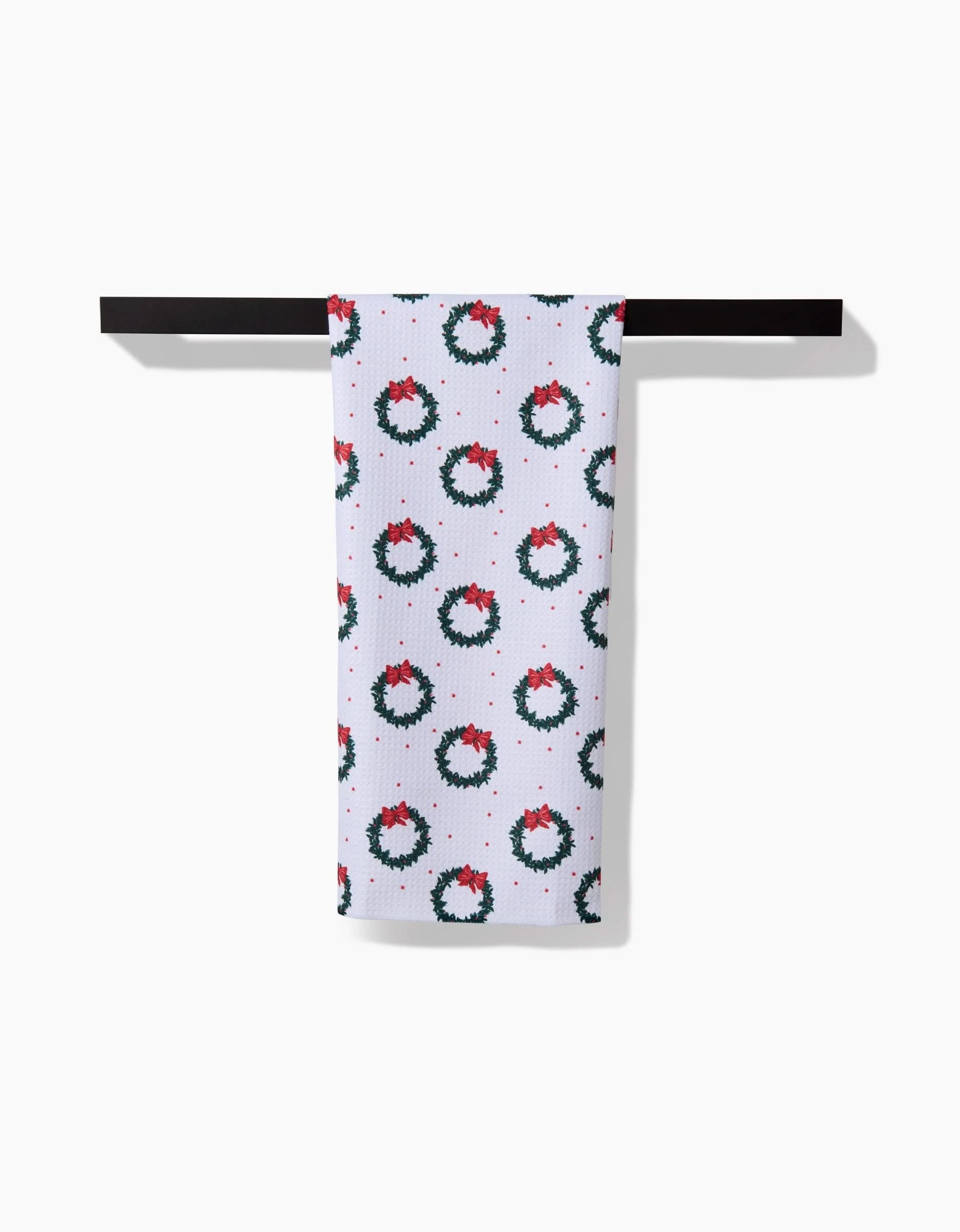 The Ash Cascade Bow Wreath Luxe Hand Towel features a repeating green wreath and red berry pattern, hanging on a black bar. Made with Geoweave technology, it offers softness and quick-drying performance.