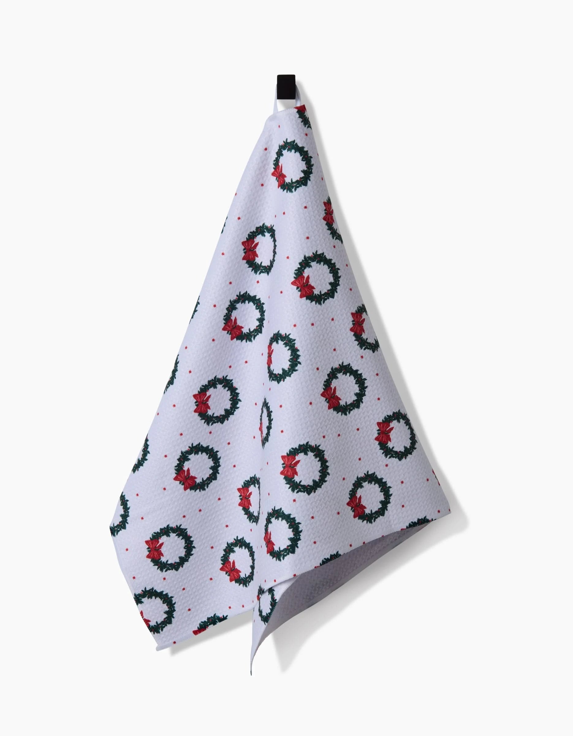 A white Ash Cascade Bow Wreath Luxe Hand Towel hangs from a black hook, showcasing green Christmas wreaths with red bows and small red dots on a white background.