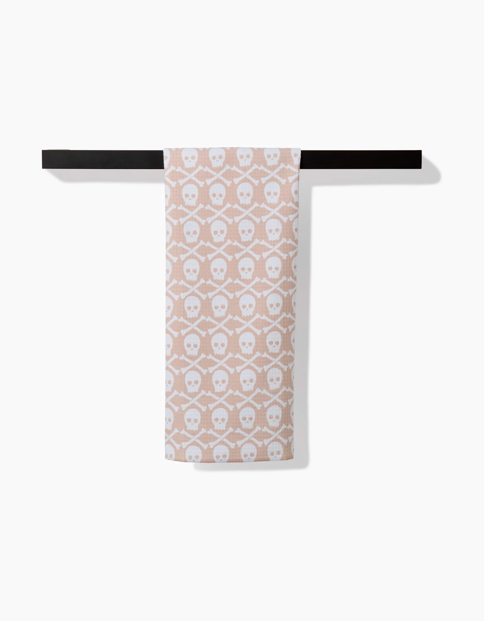 A Hufton Studio Skull Bones Natural hand towel, featuring a beige hue with white skull motifs and ultra-absorbent Geoweave fabric, hangs neatly folded on a black bar against a plain white background.