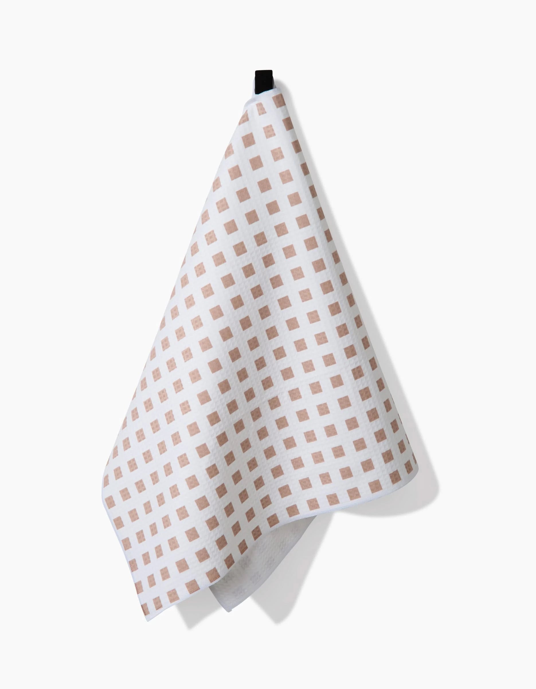 The GEOMETRY Spring Square towel, featuring light brown squares on white and crafted with ultra-absorbent Geoweave technology, hangs from a black loop against a plain white background.