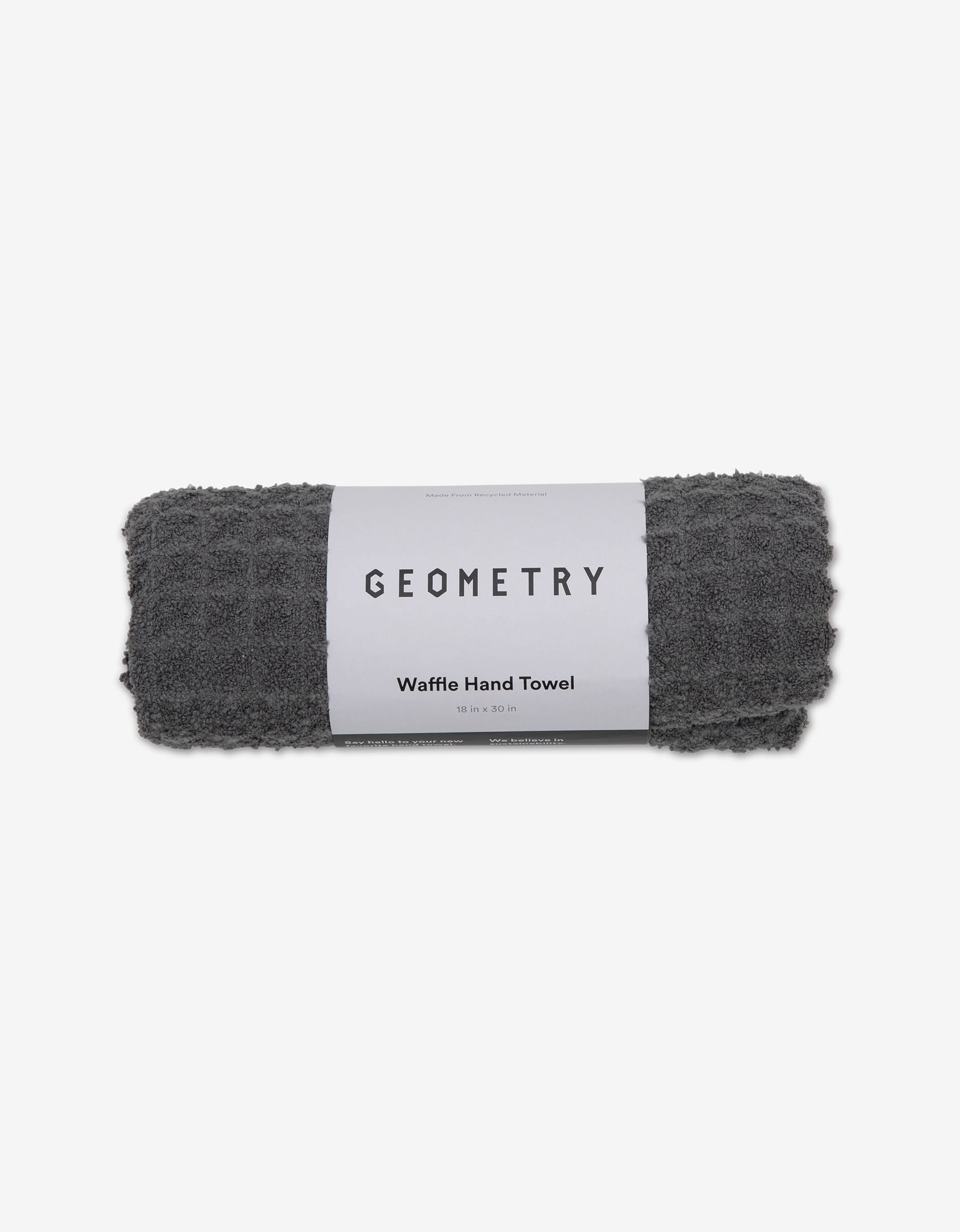 A dark gray, waffle-textured Charcoal hand towel by GEOMETRY is rolled up with a white label reading GEOMETRY, displayed against a plain white background.