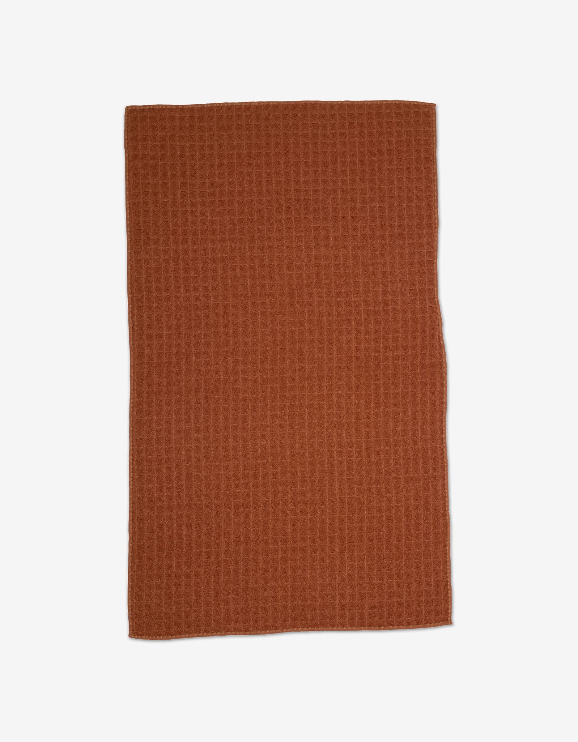A rectangular, rust-colored GEOMETRY Clay Waffle Hand Towel with a textured finish is laid flat on a white background.