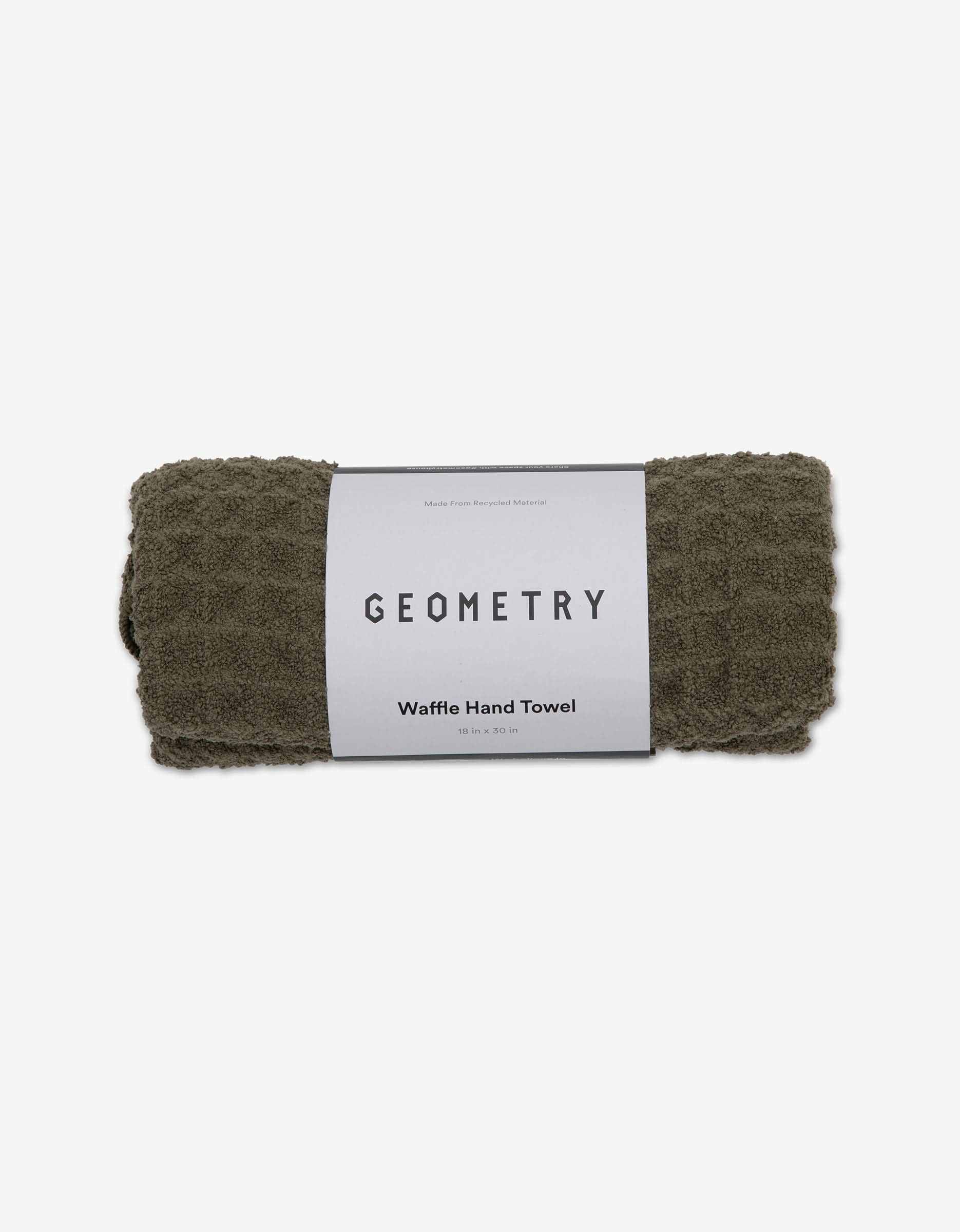 A rolled dark olive green GEOMETRY Waffle Hand Towel in Forest with a white label is displayed on a plain white background, highlighting this eco-friendly towel’s waffle texture.