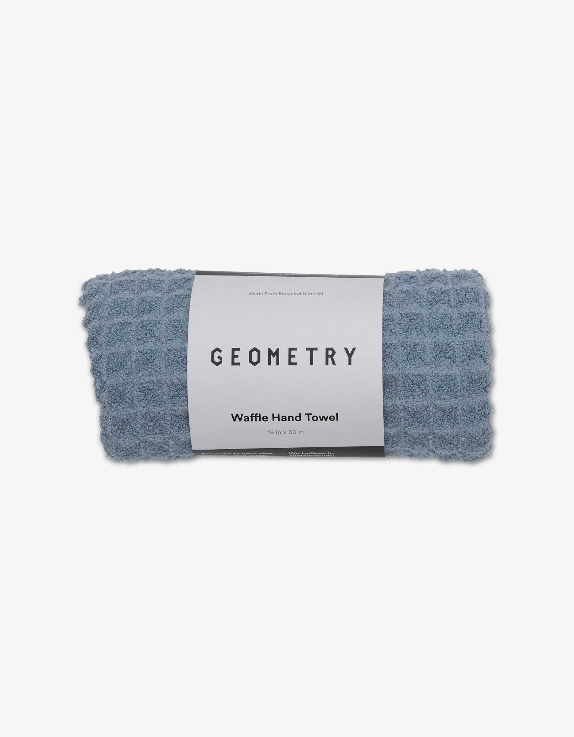 A rolled blue GEOMETRY Waffle Hand Towel called Sky, with a textured pattern and white packaging. Made from recycled material, this ultra-absorbent towel measures 19 x 29 inches.