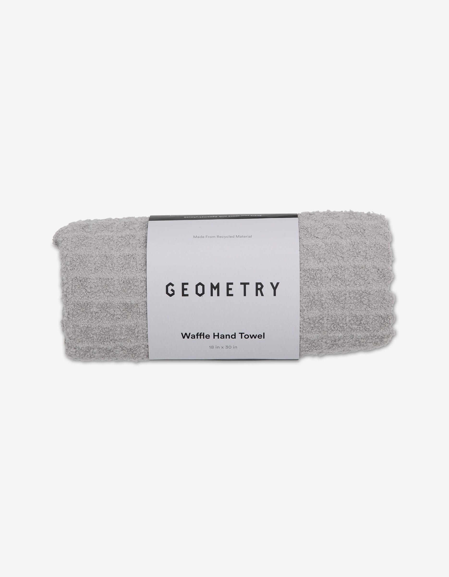 A rolled-up light gray GEOMETRY Waffle Hand Towel in the Stone color, featuring a textured pattern and wrapped in a branded label. This ultra-absorbent towel adds luxury to any space. The background is plain white.