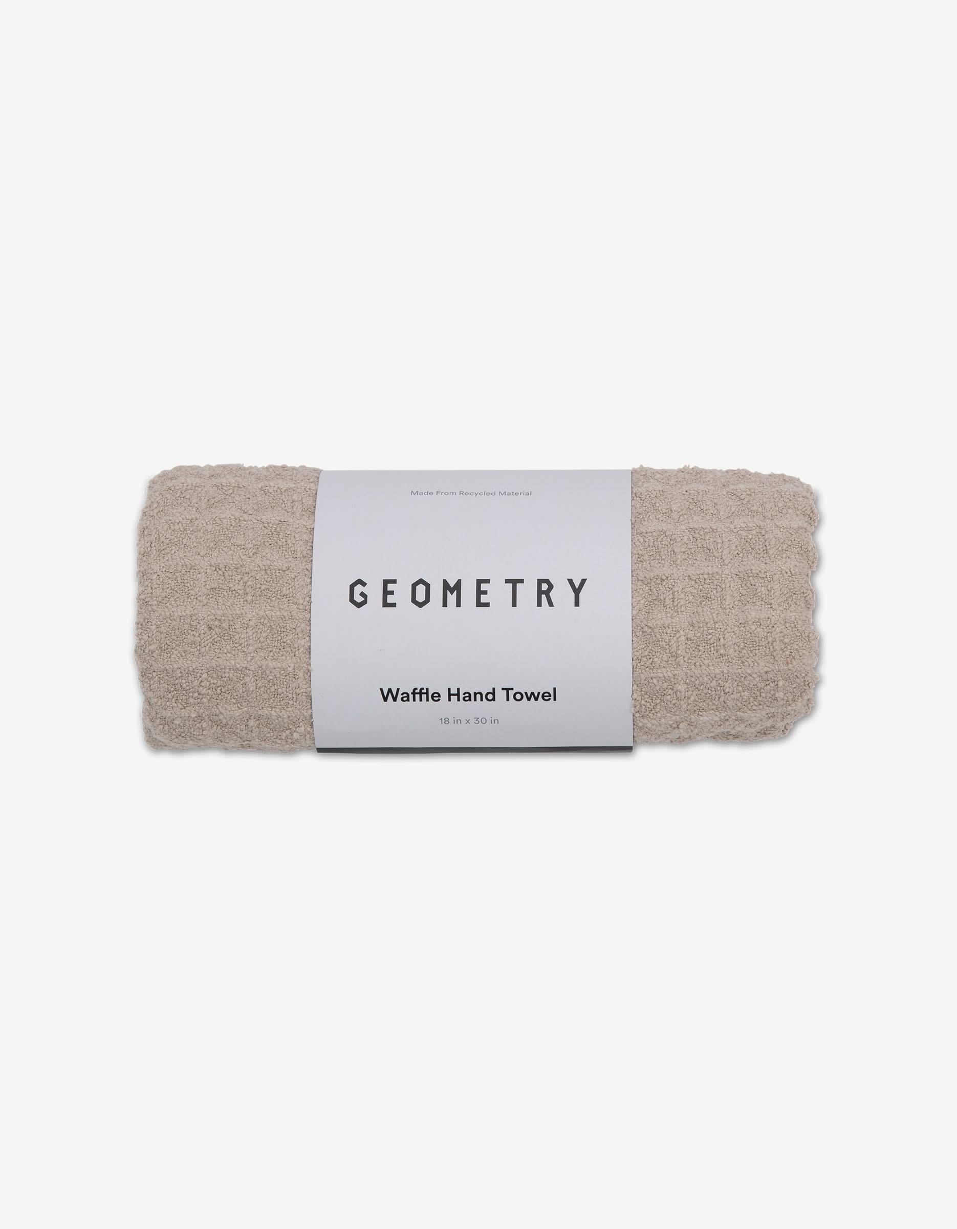 A rolled taupe GEOMETRY Waffle Hand Towel, made from recycled materials with a white label, is displayed against a white background.