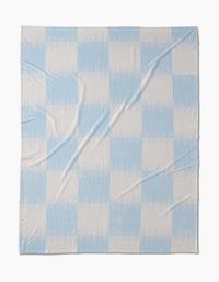 The Sweet Checks blanket by Geometry features a large, simple checkered pattern in pale blue and off-white. Made from recycled materials, its soft fabric with subtle wrinkles offers a cozy, sustainable bedding choice.