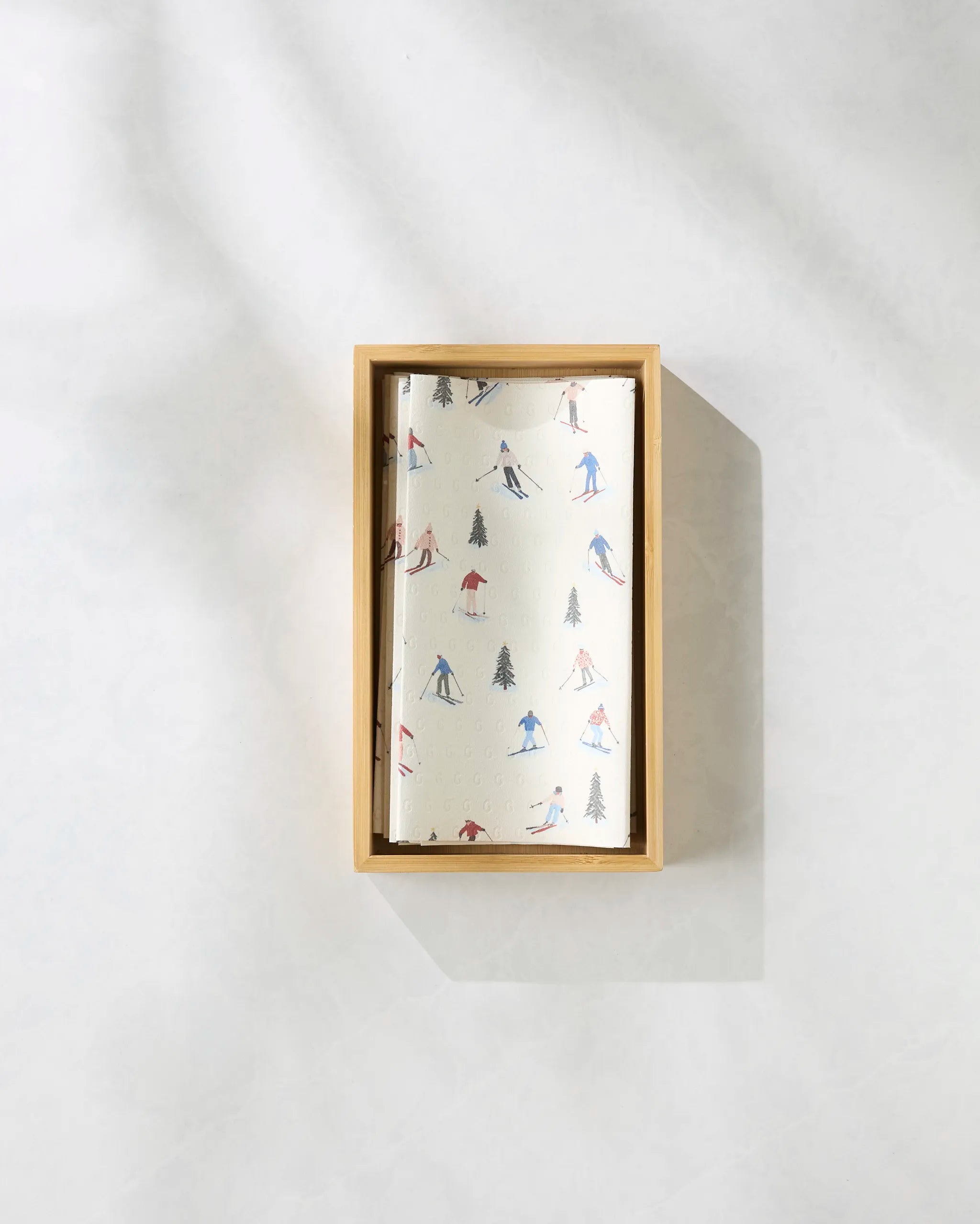 A top-down view of Geometry’s Alpine Run rectangular wooden box, filled with folded reusable paper towels featuring a small skiers and trees pattern, on a light, shadowed surface.