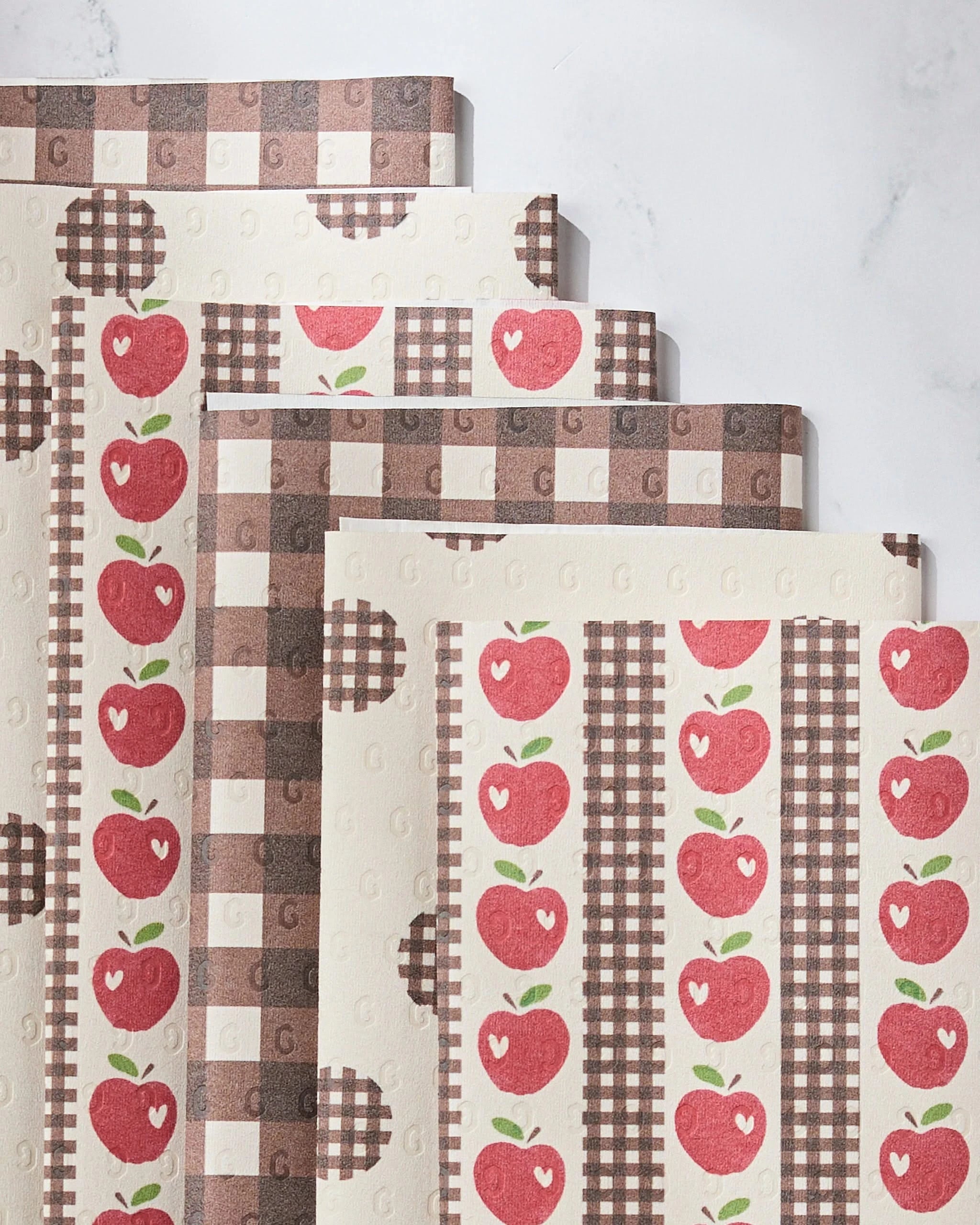 Four Geometry Apple of My Eyes Not Paper Towels are layered on a white surface, showcasing red apple prints, brown gingham checks, and stripes for a rustic, country-inspired touch in your eco-friendly kitchen.
