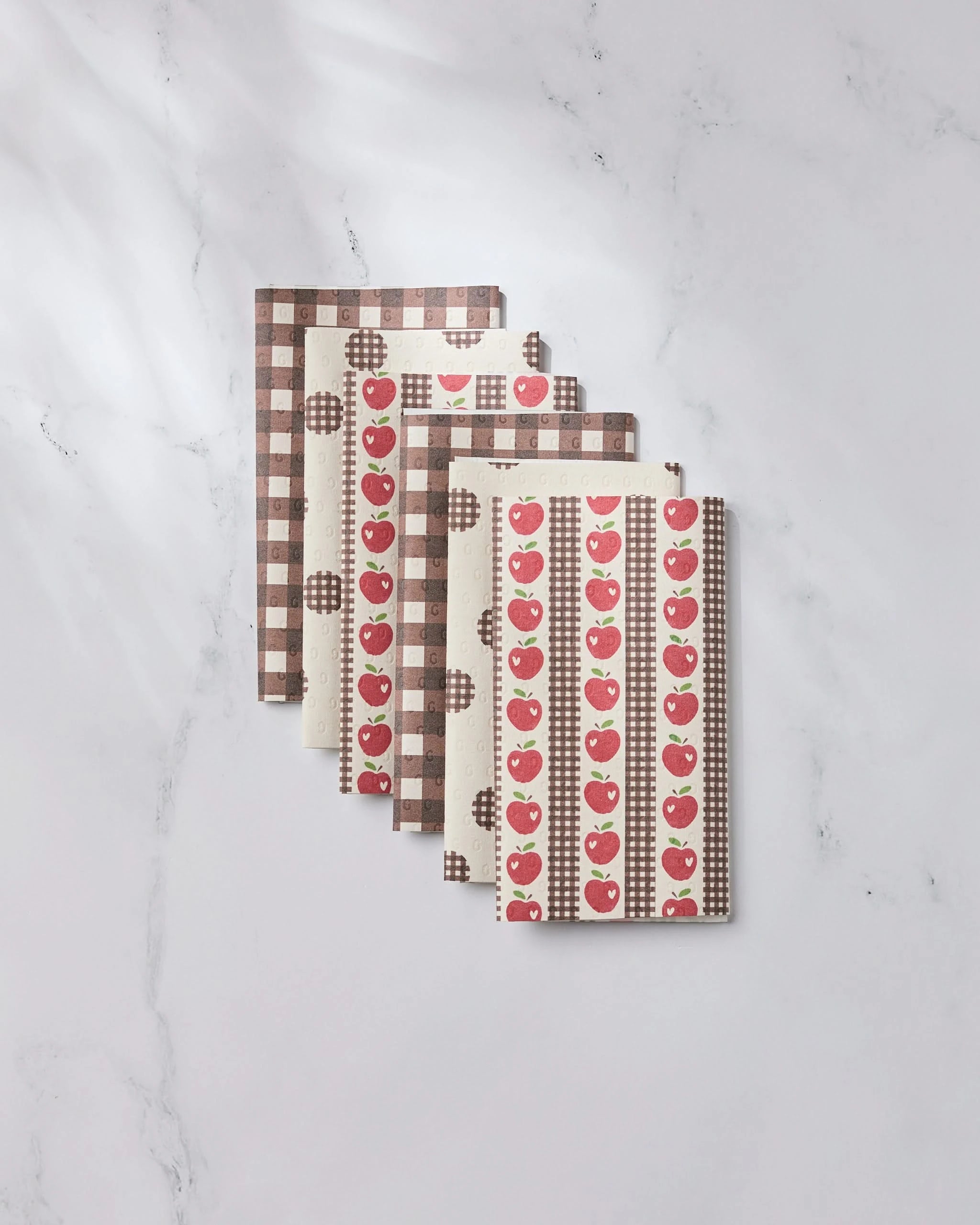 Five Apple of My Eyes notebooks by Geometry are arranged in a fan shape on white marble, featuring eco-friendly kitchen towel-inspired designs with red apples and brown-and-white checkered patterns.
