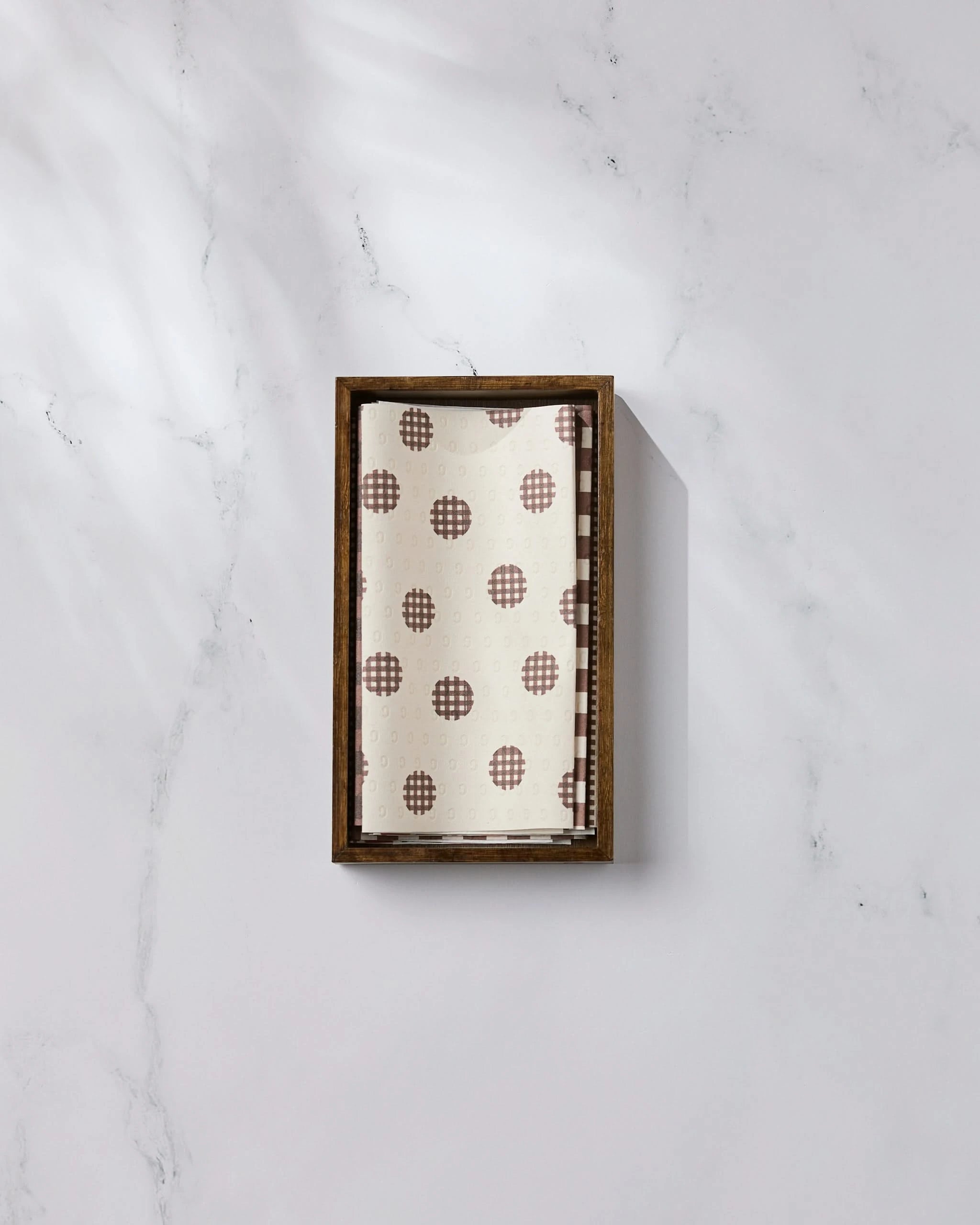 The Apple of My Eyes kitchen towel by Geometry, featuring a brown checkered circle design on white, is neatly folded in a rectangular wooden tray atop a white marble surface with subtle veins and soft shadows.