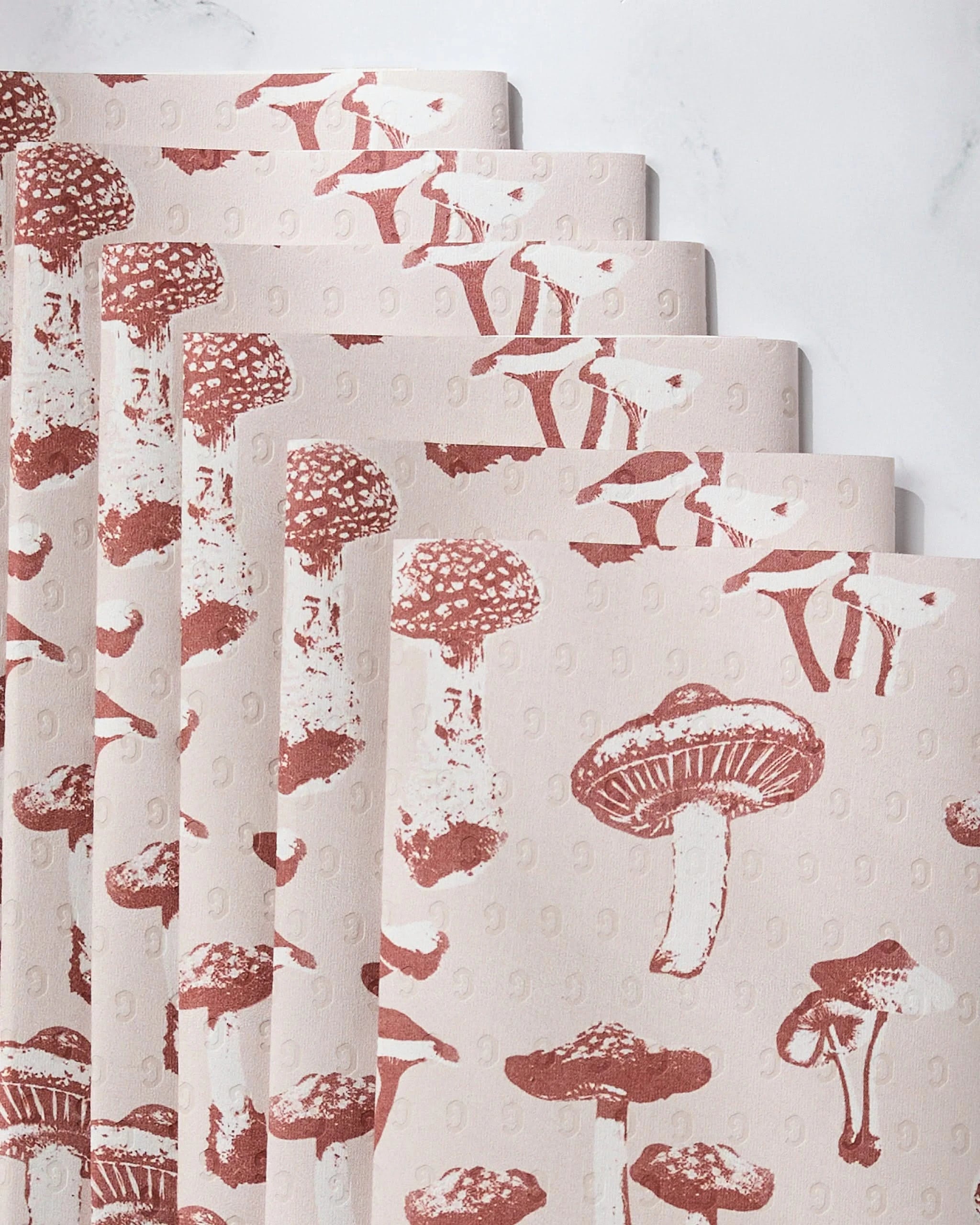 A fanned stack of Geometry’s Autumn Mushrooms Not Paper Towels, featuring a repeating red mushroom pattern, displayed on light marble—an eco-friendly and stylish addition to any kitchen.
