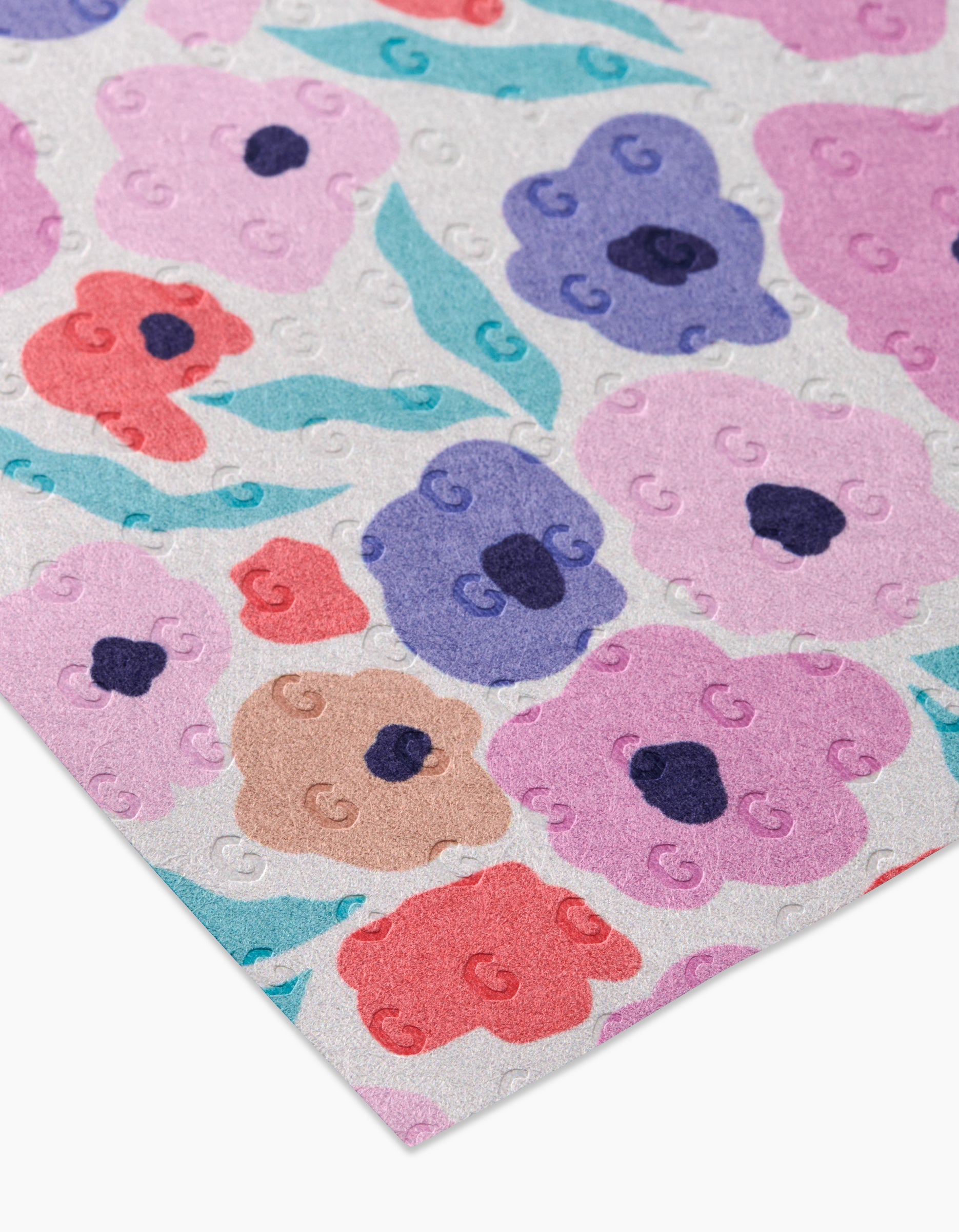 A close-up of GEOMETRYs Blossoming Love shows a sheet of embossed, post-consumer recycled paper with pink, purple, red, and blue flowers plus green leaves on a light background.