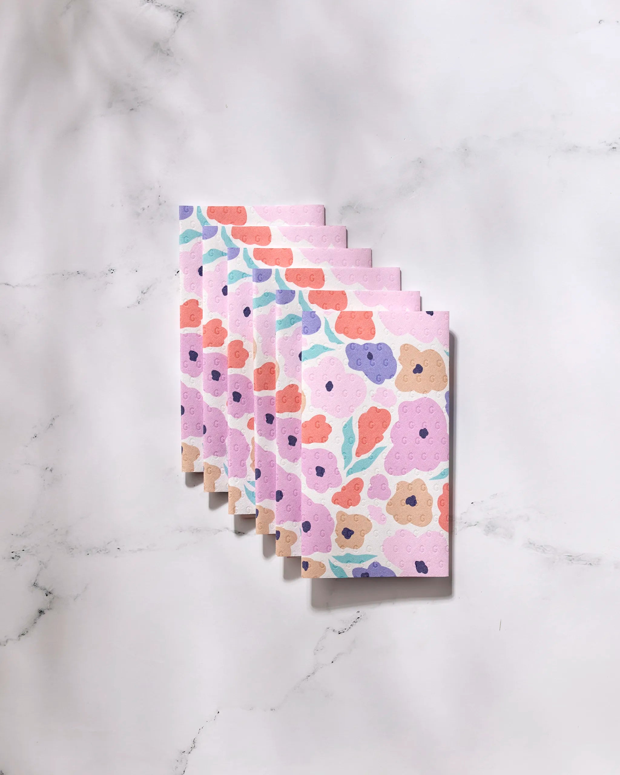 Six "Blossoming Love" GEOMETRY reusable towels, in pastel abstract floral patterns of pink, purple, orange, and green, are fanned out on white marble—offering an eco-friendly alternative to Not Paper Towels.
