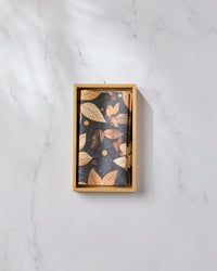 A wooden box of Geometry’s Botanical Wonderland cards, featuring gold leaves and dots on black, rests on white marble next to eco-friendly kitchen towels, all bathed in soft shadows.