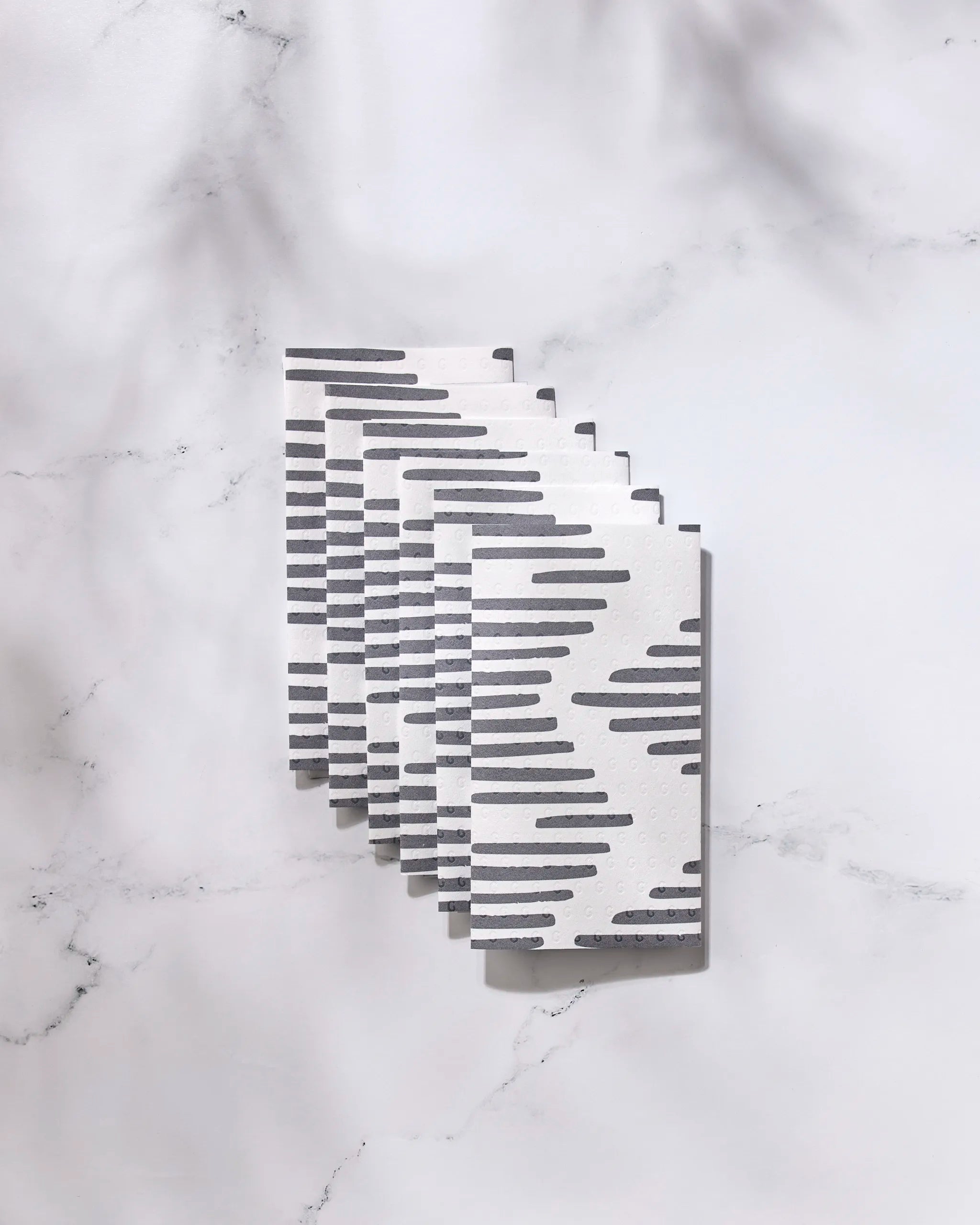 Six Charlie textured cards by GEOMETRY, featuring horizontal black stripes, are fanned out on a light marble surface and evoke the look of eco-friendly kitchen towels, with soft shadows above.