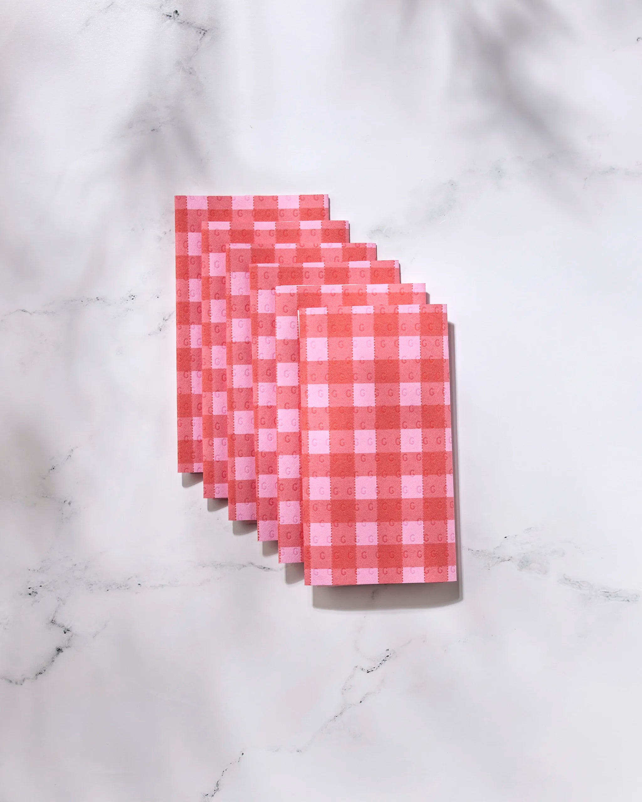 Seven Cherry Gingham reusable paper towels by GEOMETRY, featuring a pink and red gingham pattern, are neatly arranged in a row on a light marble surface with soft shadows.