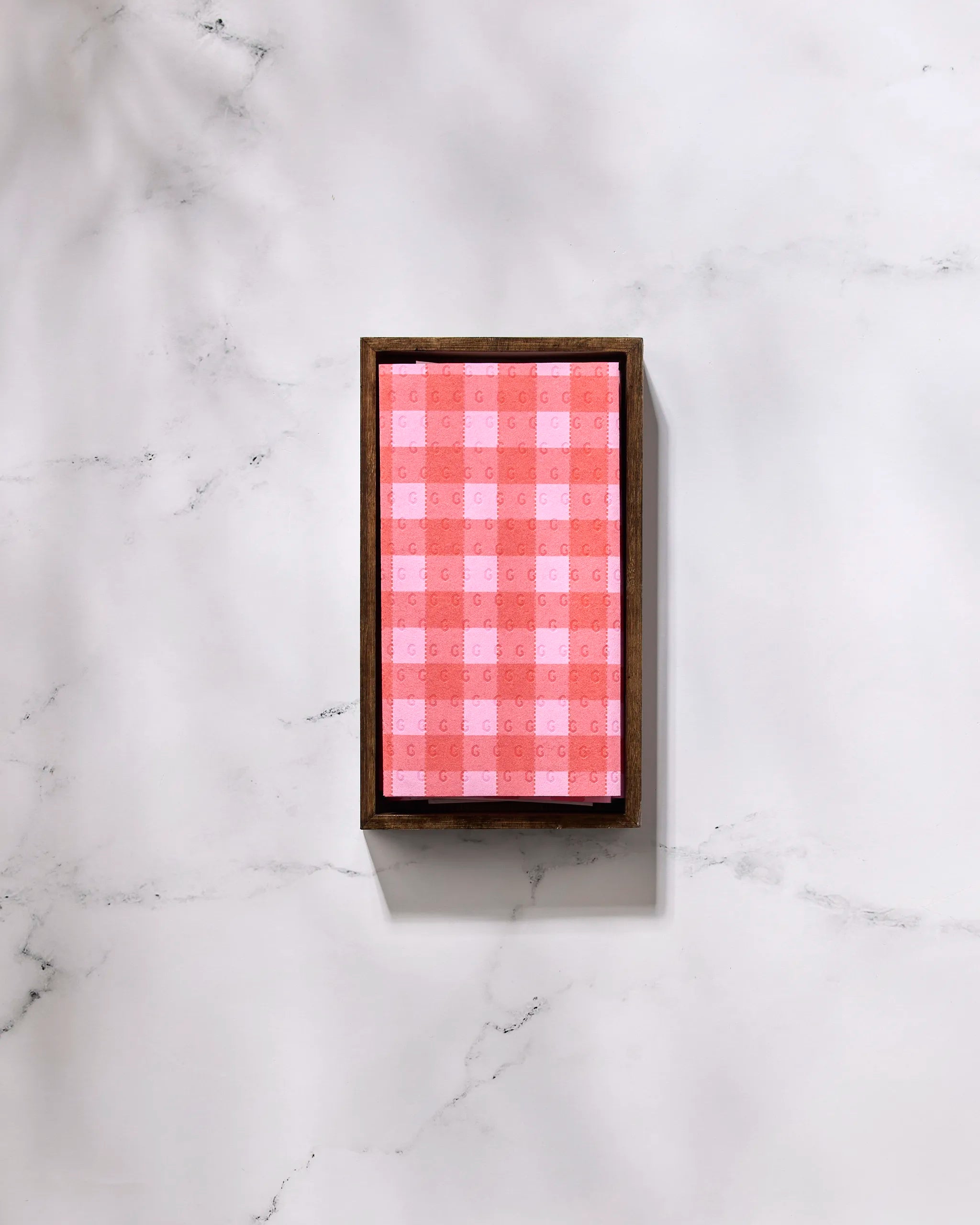 A box of GEOMETRY Cherry Gingham eco-friendly kitchen towels, featuring a red and pink checkered pattern and framed by a thin dark brown border, rests on a white marble surface.