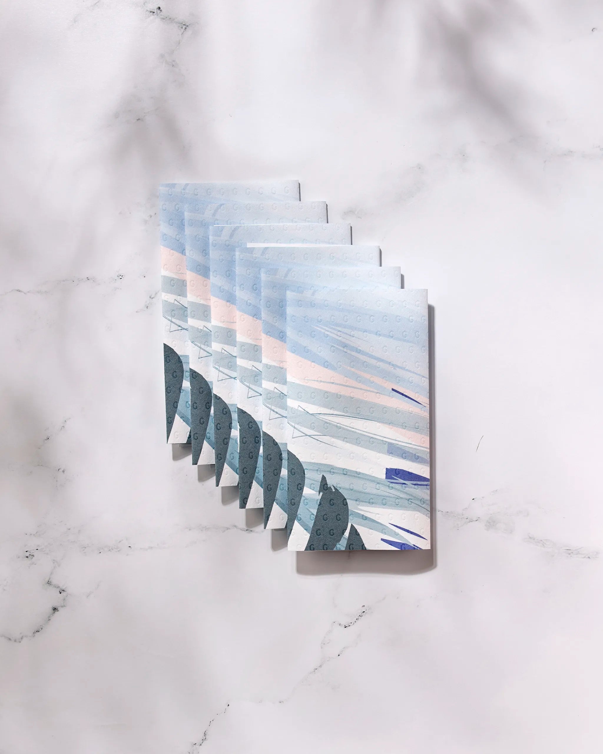 Seven “Create” booklets by GEOMETRY, featuring abstract blue, pink, and gray covers, are fanned out on white marble—suggesting how eco-friendly kitchen towels can stylishly complement your sustainable home. Light shadows fall in the upper right corner.