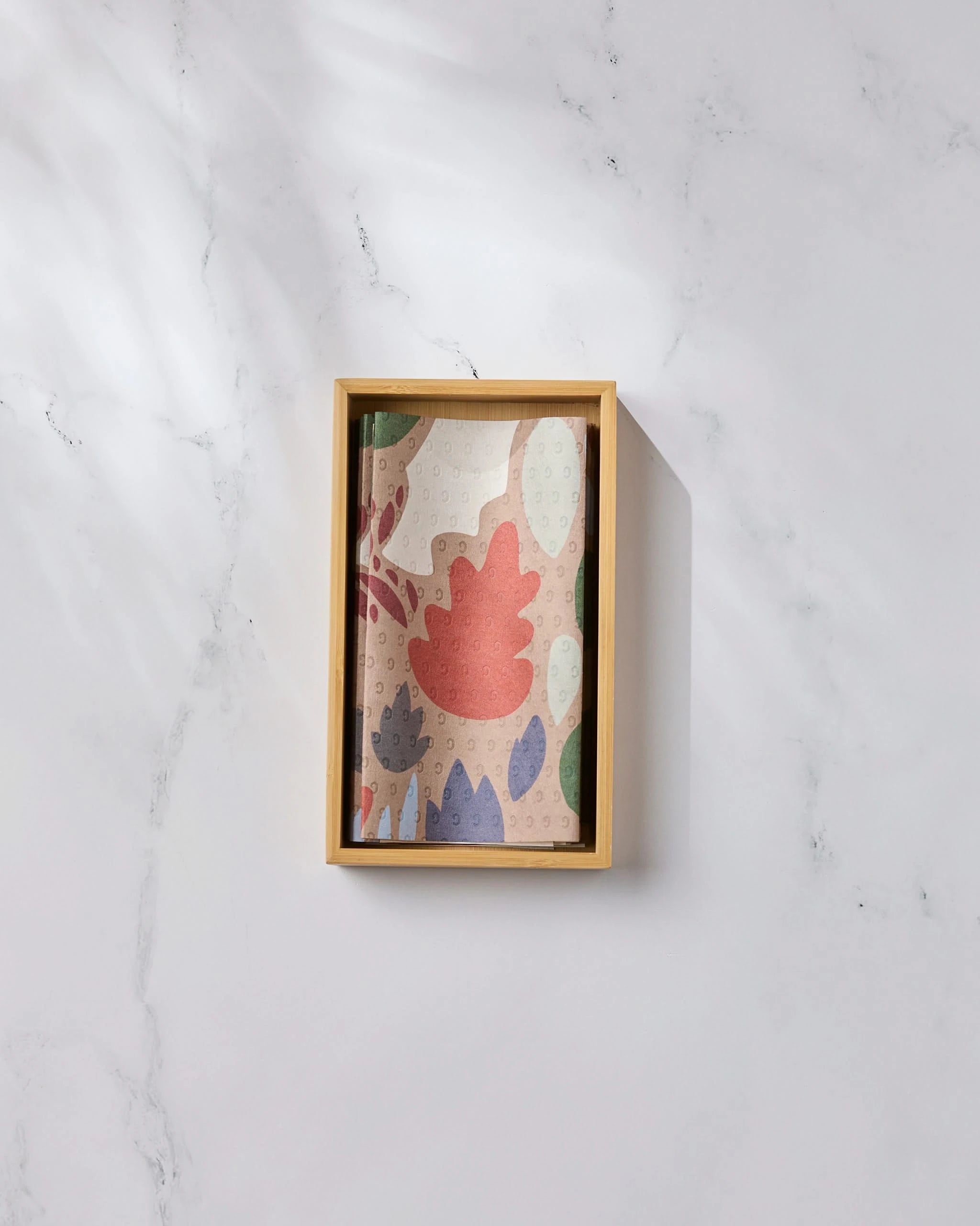 The Geometry Cut Paper Leaves Mocha notebook, featuring a colorful abstract leaf design made from post-consumer recycled materials, rests in a wooden tray on white marble, bathed in soft natural light.