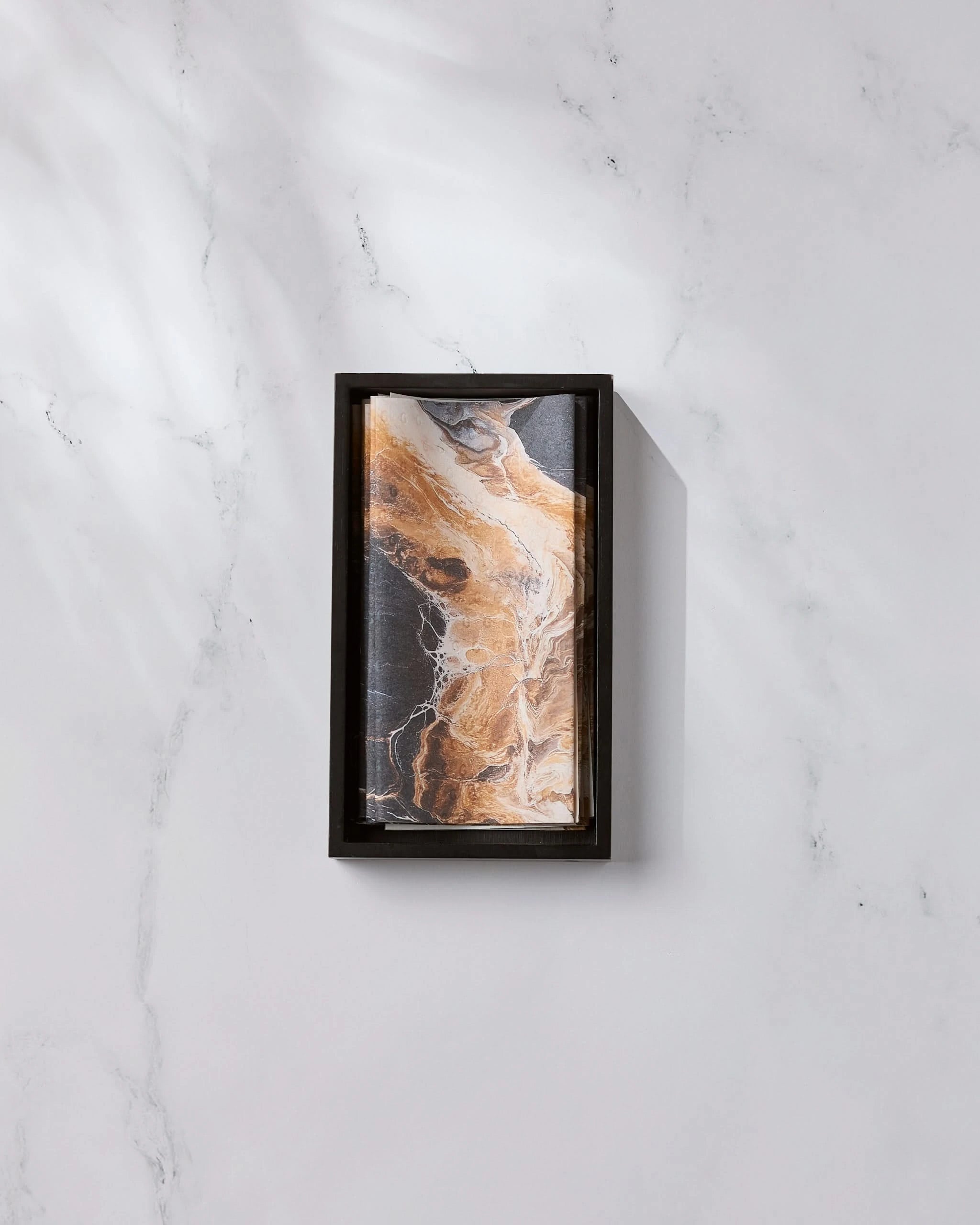 The Emperador by Geometry, featuring a rectangular black frame and an abstract brown, black, and white marble-inspired pattern, rests on a white marble surface with light gray veining as diagonal shadows fall across the scene.