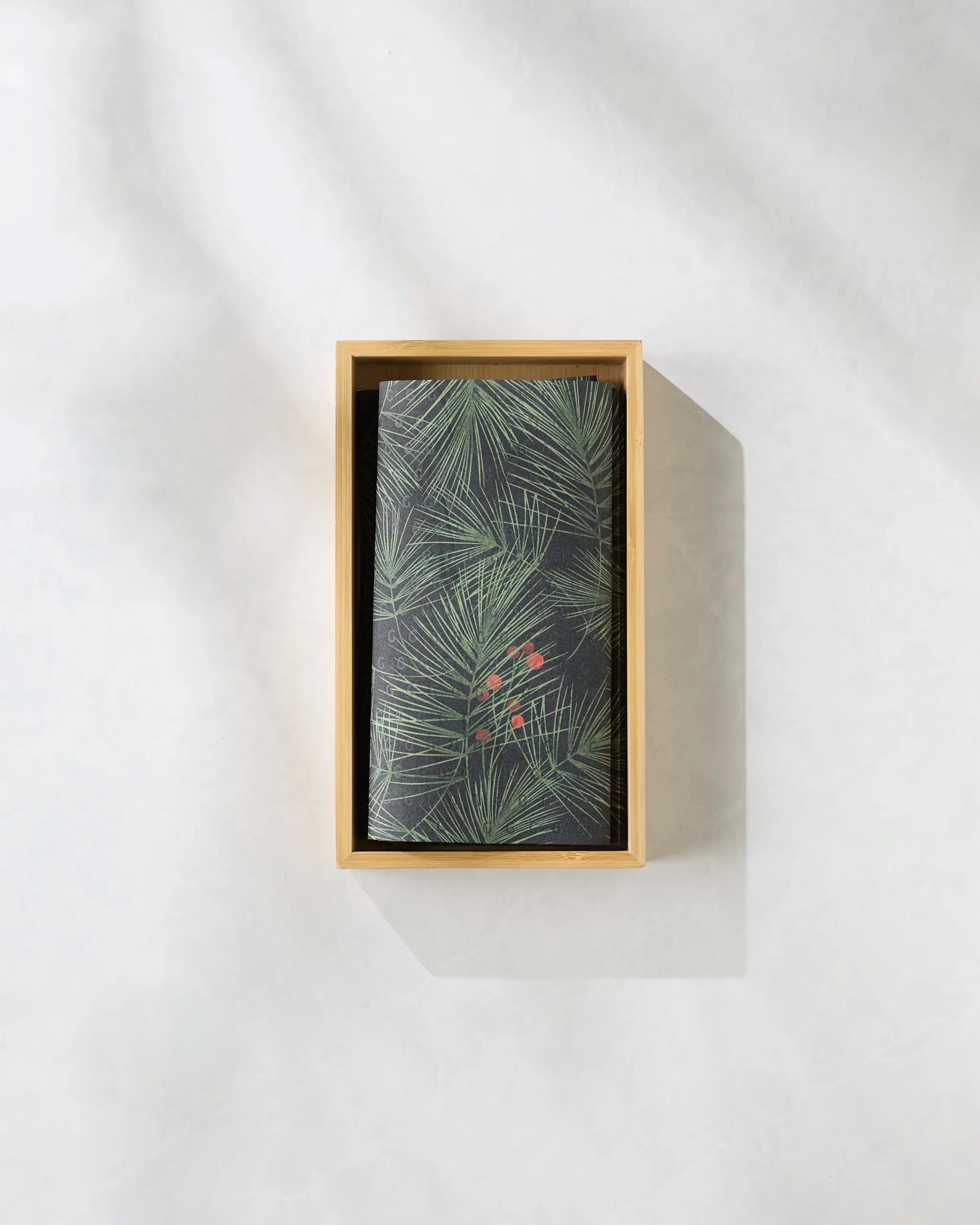 The Geometry Evergreen Pine notebook with a dark cover of green pine needles and red berries rests in a light wooden box on a gray surface—an ideal match with eco-friendly paper towels for a sustainable workspace.