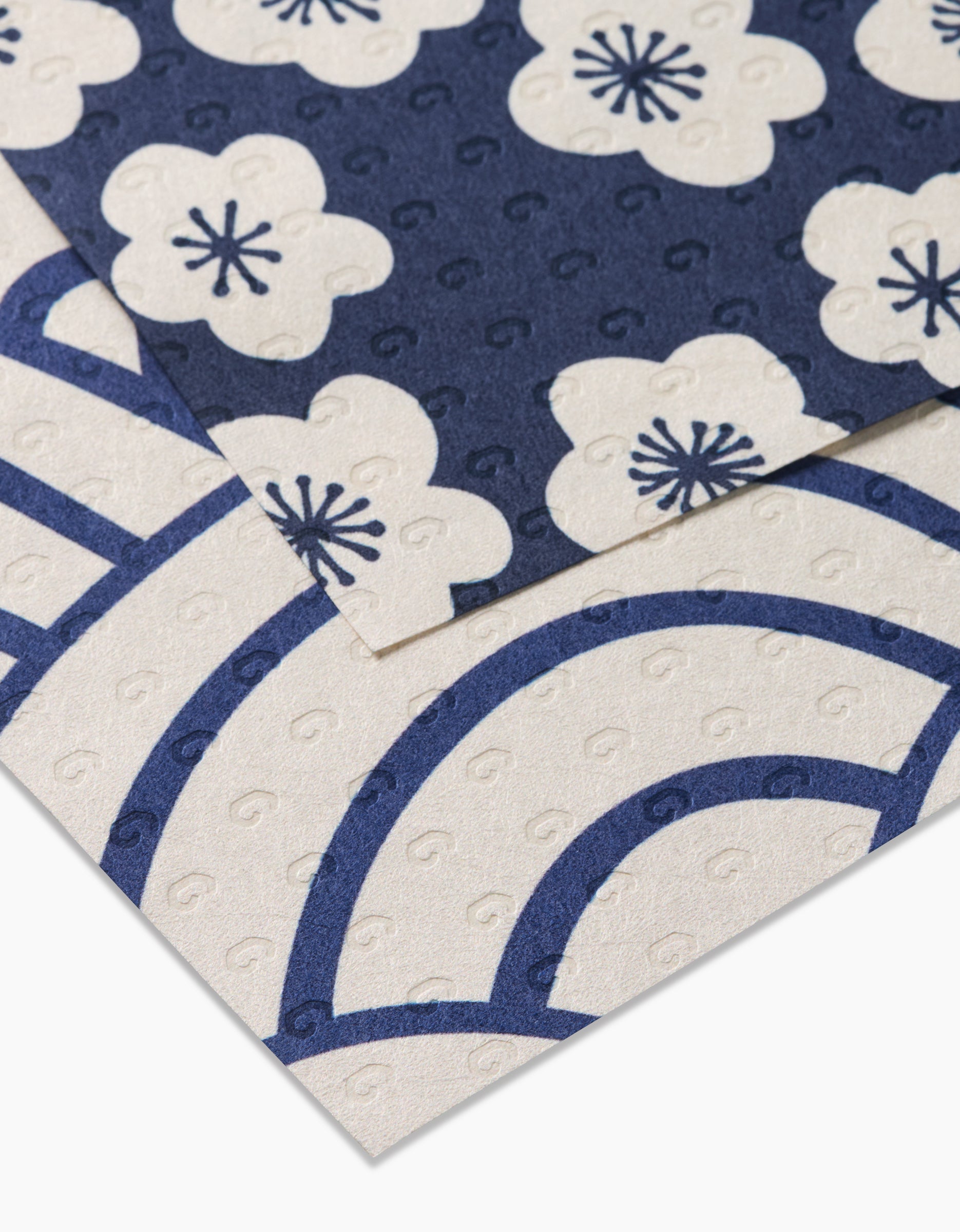 Two Every Wish sheets by GEOMETRY overlap: the top features navy blue with white plum blossoms, while the bottom shows a white base with bold blue arches and swirls. Both are reusable and textured.