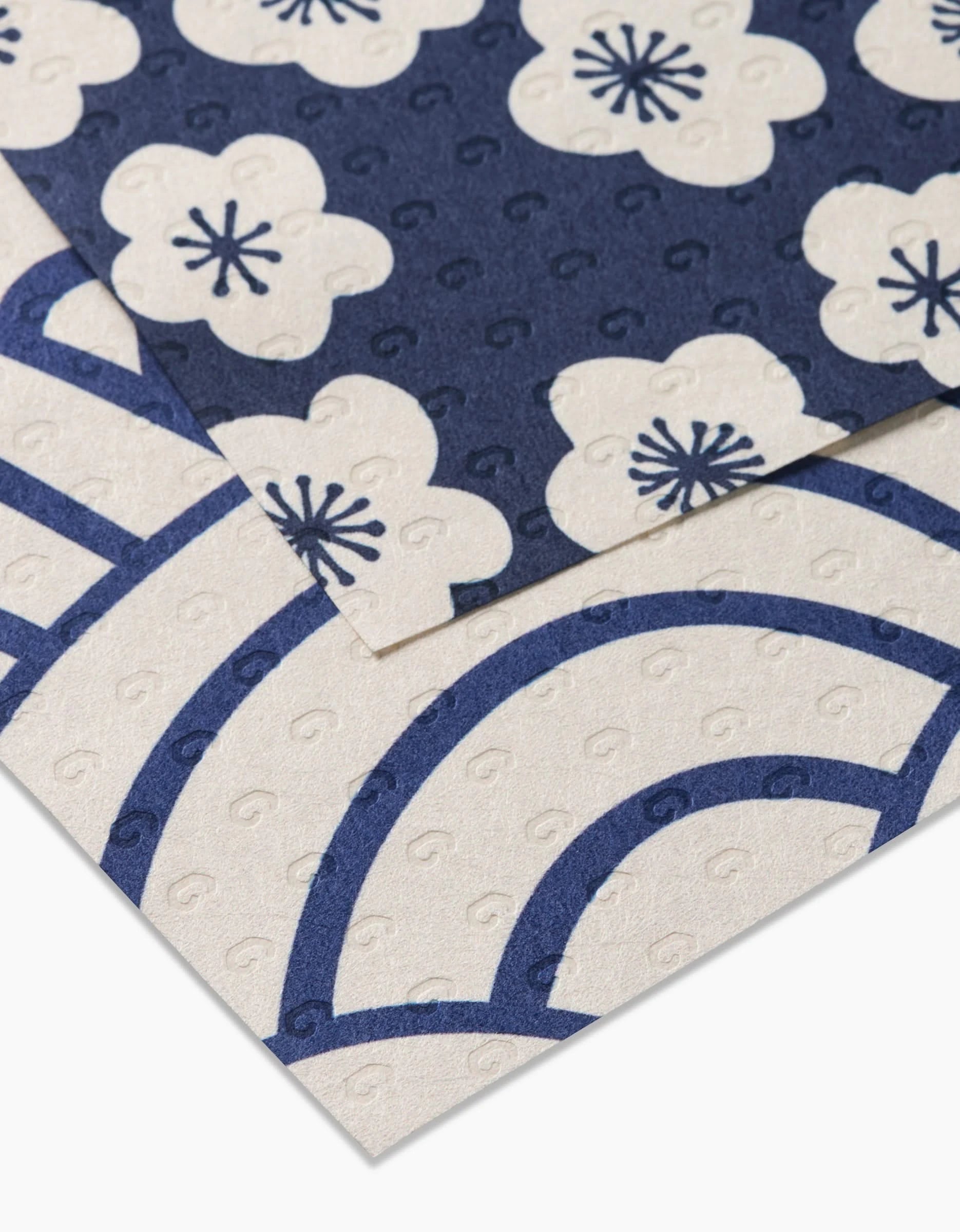 Two Every Wish sheets by GEOMETRY overlap: the top features navy blue with white plum blossoms, while the bottom shows a white base with bold blue arches and swirls. Both are reusable and textured.