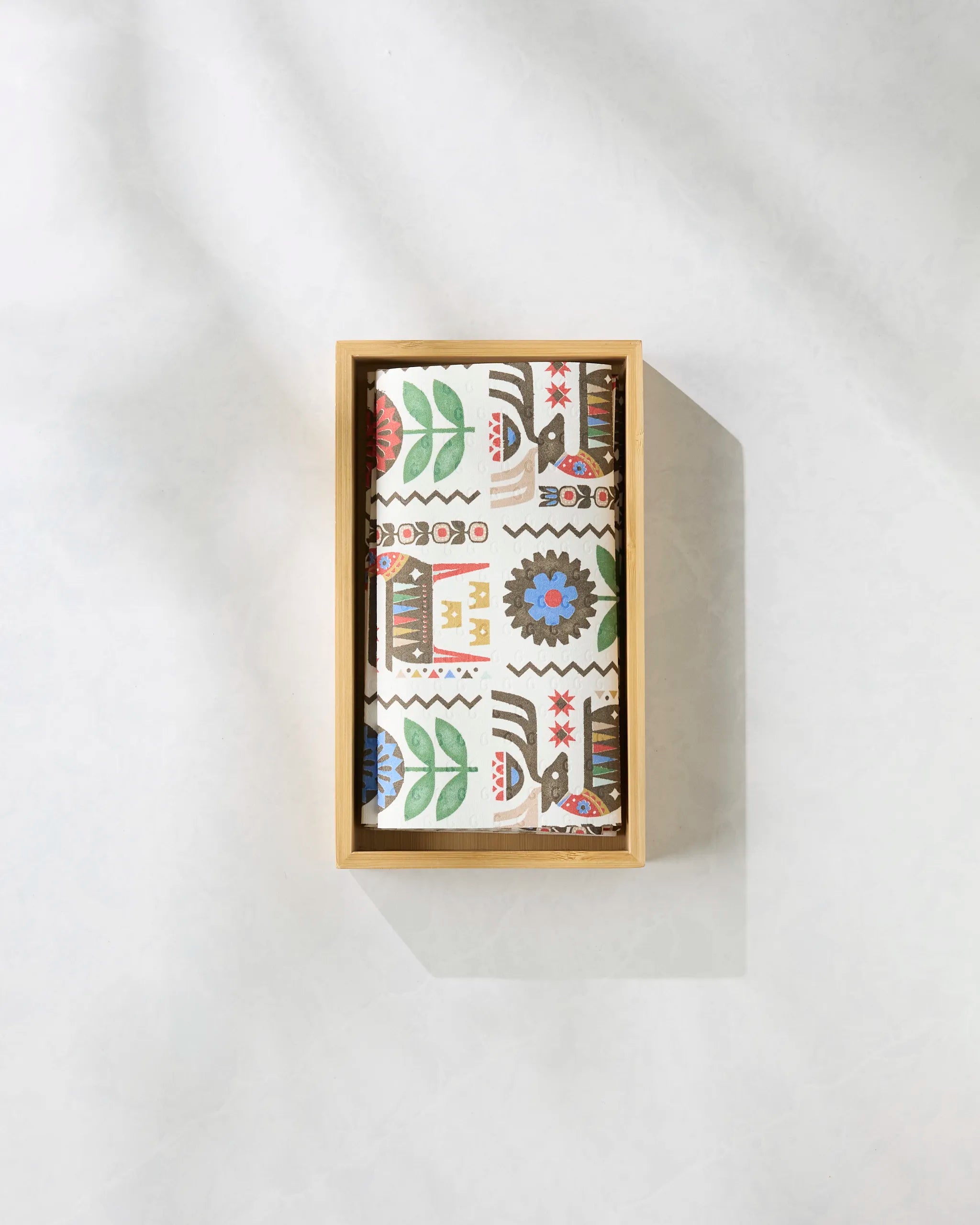 The Folk Deer notebook by Geometry features a vibrant, folk-inspired floral and geometric pattern and eco-friendly pages, presented in a light wooden box on a sunlit surface.