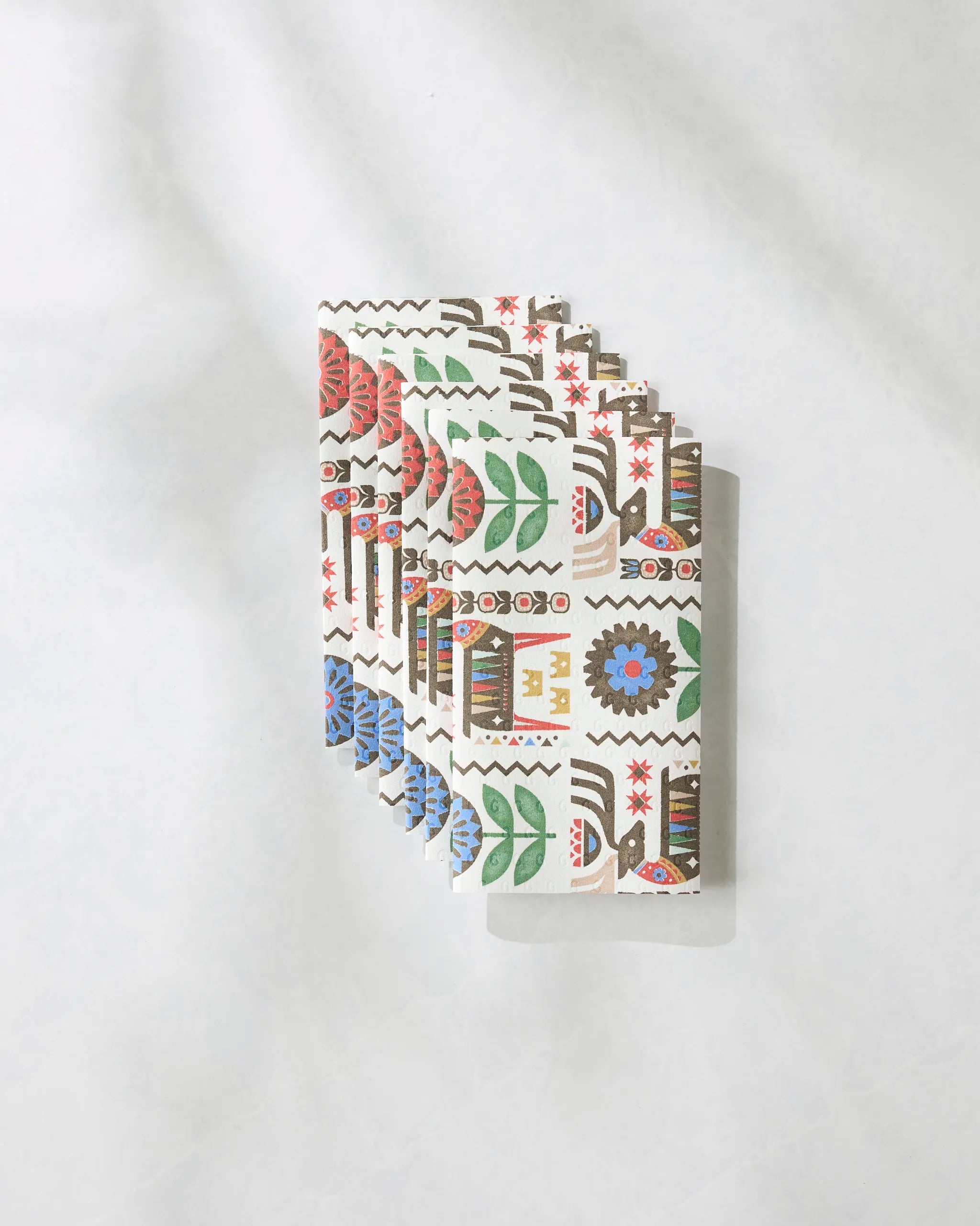 Five folded Geometry reusable paper towels in the Folk Deer design, featuring colorful, folk-inspired patterns with flowers, leaves, and geometric shapes, are stacked on a light, textured background.