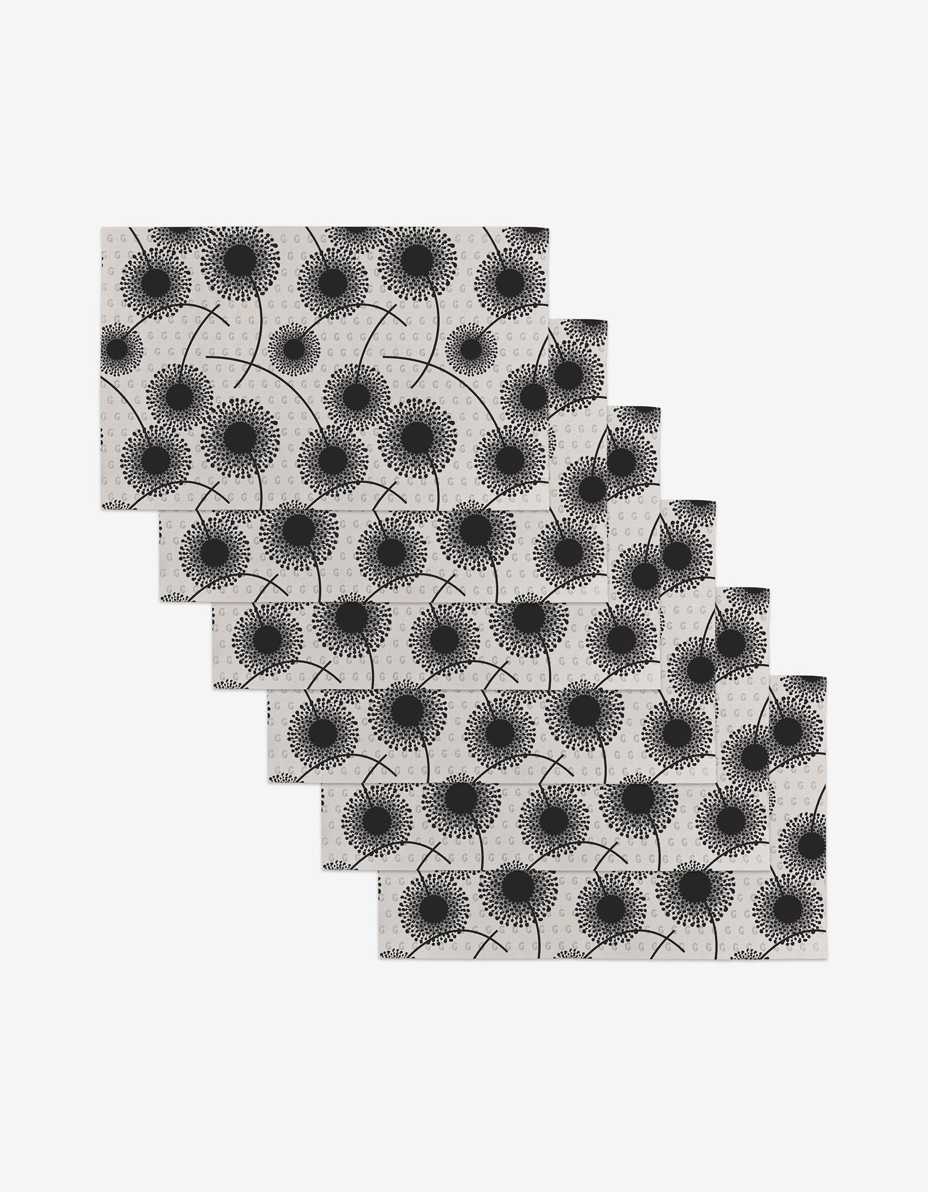 Six Fully Bloomed rectangular reusable placemats, featuring a black dandelion pattern on light gray, are arranged in a cascading diagonal stack on a white surface.