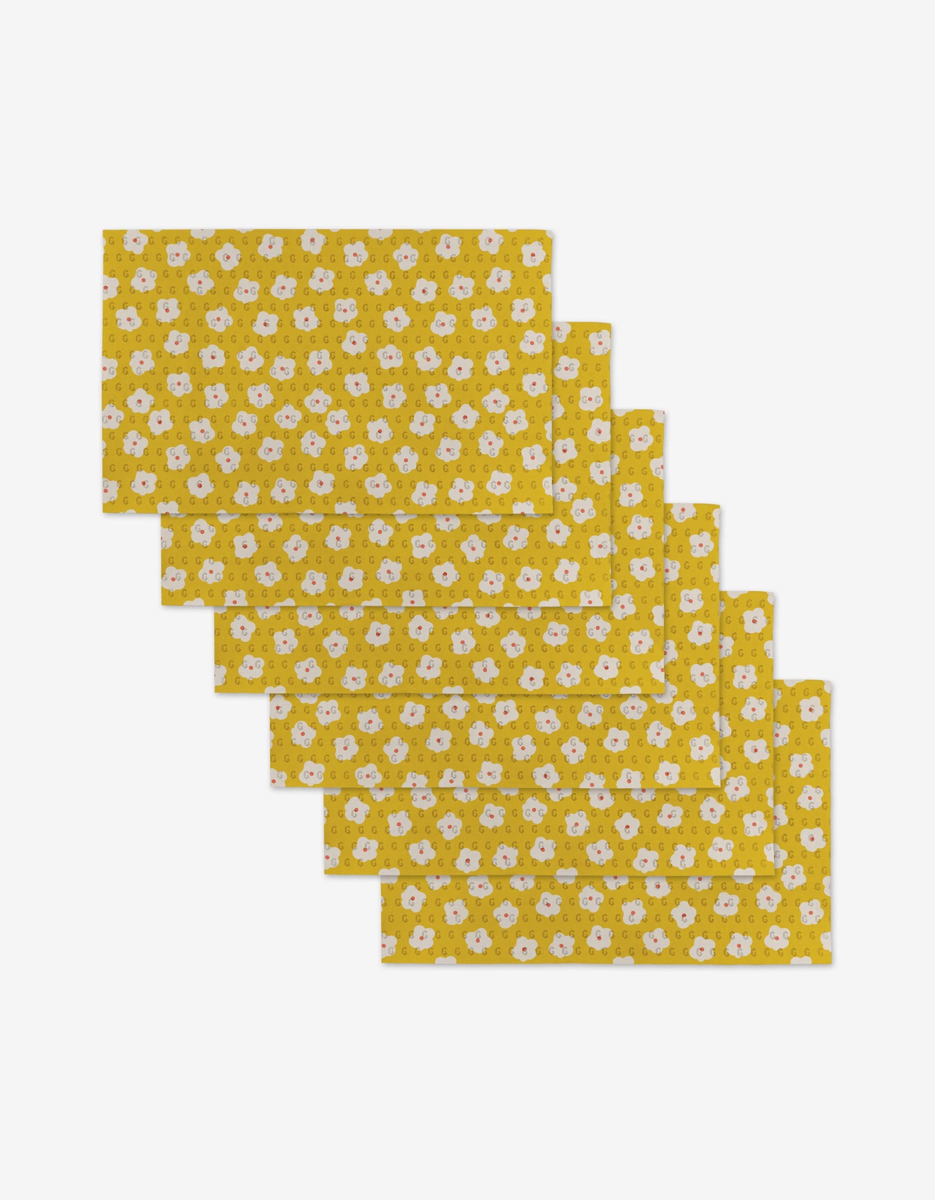 Six Sara Mari Garden Crush placemats, rectangular and yellow with a floral pattern of white flowers and green leaves, made from eco-friendly materials, are stacked in a slightly overlapping arrangement on a white background.