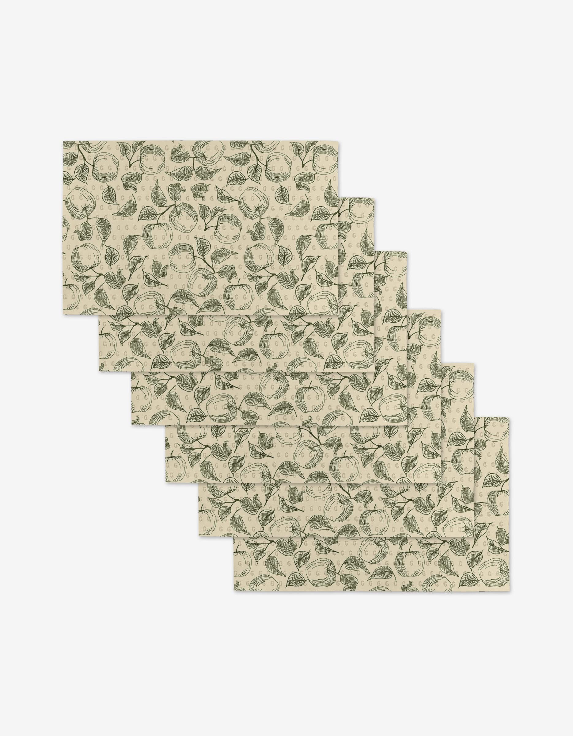 Five rectangular Gravenstein Apple placemats by Ash Cascade, beige with green botanical prints of leaves and fruit, are arranged in a staggered layout on a white background—an eco-friendly alternative to traditional placemats.