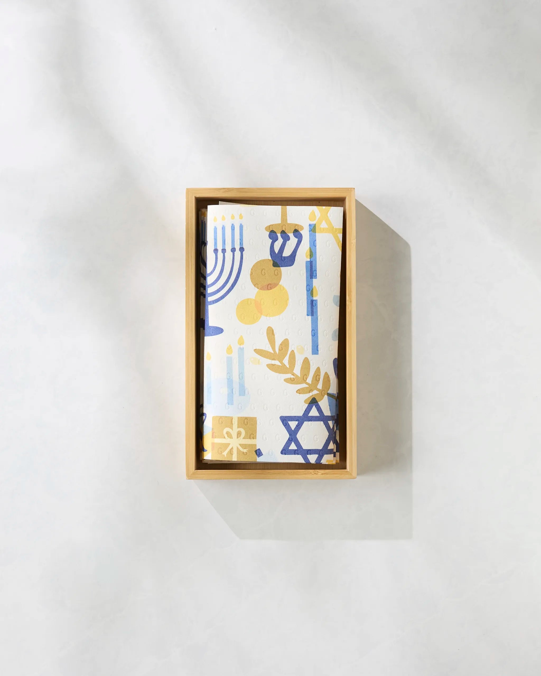 The "Hanukkah is Here" box by Geometry includes eco-friendly paper towels with menorahs, Stars of David, and dreidels in blue, gold, and white on a light background—perfect for a sustainable Hanukkah celebration.